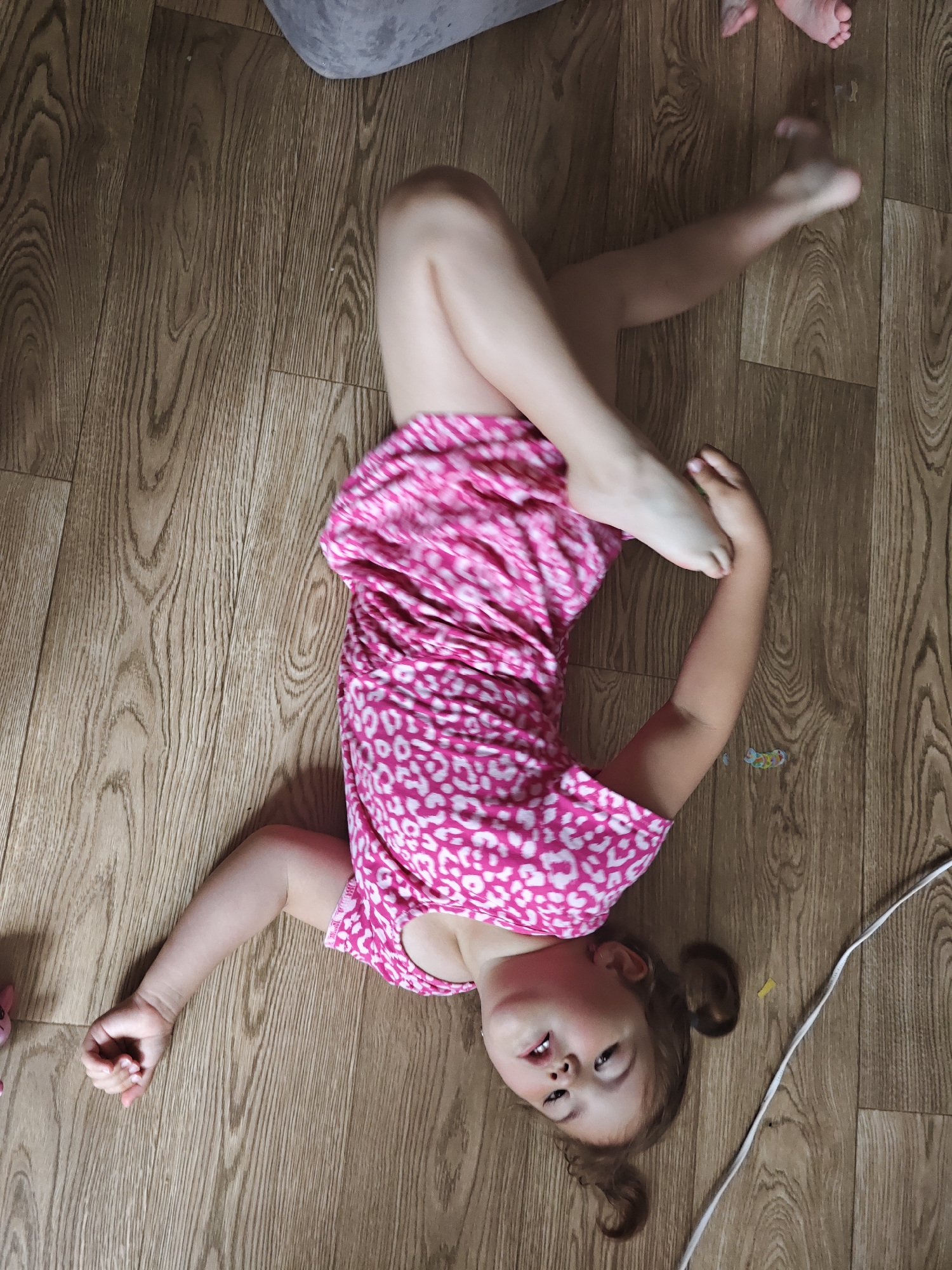The image shows a young girl lying on a wooden floor, possibly in a home in Chaikovsky, Russia. She is wearing a pink dress with a white leopard-like print. Her body is contorted in an unusual pose, with her legs bent and crossed and her arms outstretched. Her head is tilted, and her eyes are open, looking upwards. She appears to be relaxed or playing on the floor. The lighting suggests it is daytime, and there are no indications of the weather. The wooden floor has a light brown, natural wood grain pattern. A white electrical cord runs diagonally across the lower right side of the image, and a few colorful small stickers are visible on the floor near the girl.