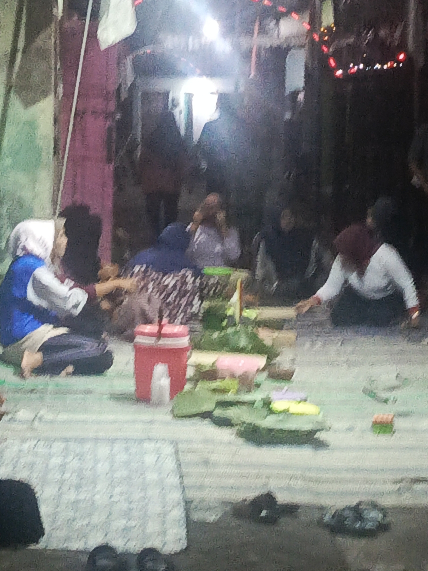 The image captures a low-angle, dimly lit scene that appears to be an outdoor gathering or event in Kediri City, Indonesia. In the foreground, a light blue and white checkered tablecloth covers a surface, with several dark objects, possibly shoes or bags, placed on the ground near it. To the left, a person wearing a blue vest over a white top and a light-colored headscarf is seated on the ground. To their right, a red plastic container with a white lid is positioned. Further into the scene, a group of people are gathered, some seated on the ground. They are participating in an activity involving food arrangements placed on what looks like banana leaves and other greenery, arranged on mats on the ground. One person, wearing a white long-sleeved shirt and a dark head covering, is reaching towards the food. The background shows a wall with a pink section and hanging red decorative lights. The overall atmosphere is casual and communal.