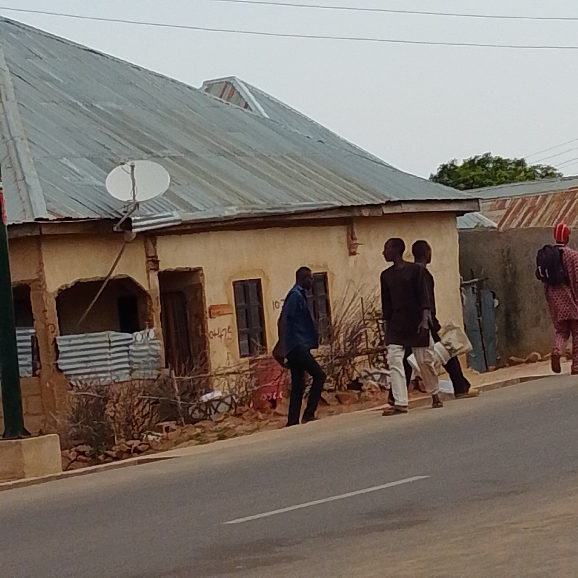 In Gombe, Nigeria, a scene unfolds on a street with a building constructed of rough, light-colored material and a corrugated metal roof. A satellite dish is affixed to the roof. The building has a recessed doorway and at least two windows, one of which is dark and appears to be covered from the inside with corrugated metal sheets. The exterior walls bear some graffiti and markings.
In front of the building, four individuals are walking on the side of the road. The closest group consists of three men walking together. One man in the foreground is wearing a dark long-sleeved shirt and light-colored pants, carrying a light-colored bag. To his left, another man in a dark shirt and light pants walks slightly behind him. A third man, also in dark clothing and light pants, is to the right of the first man, also carrying a bag. Further to the right, a fourth individual, wearing a patterned garment and a red head covering, walks away from the viewer with a backpack. The road surface is paved, with a white line visible in the lower portion of the image. The background shows more buildings with corrugated roofs and some greenery. The sky is overcast.