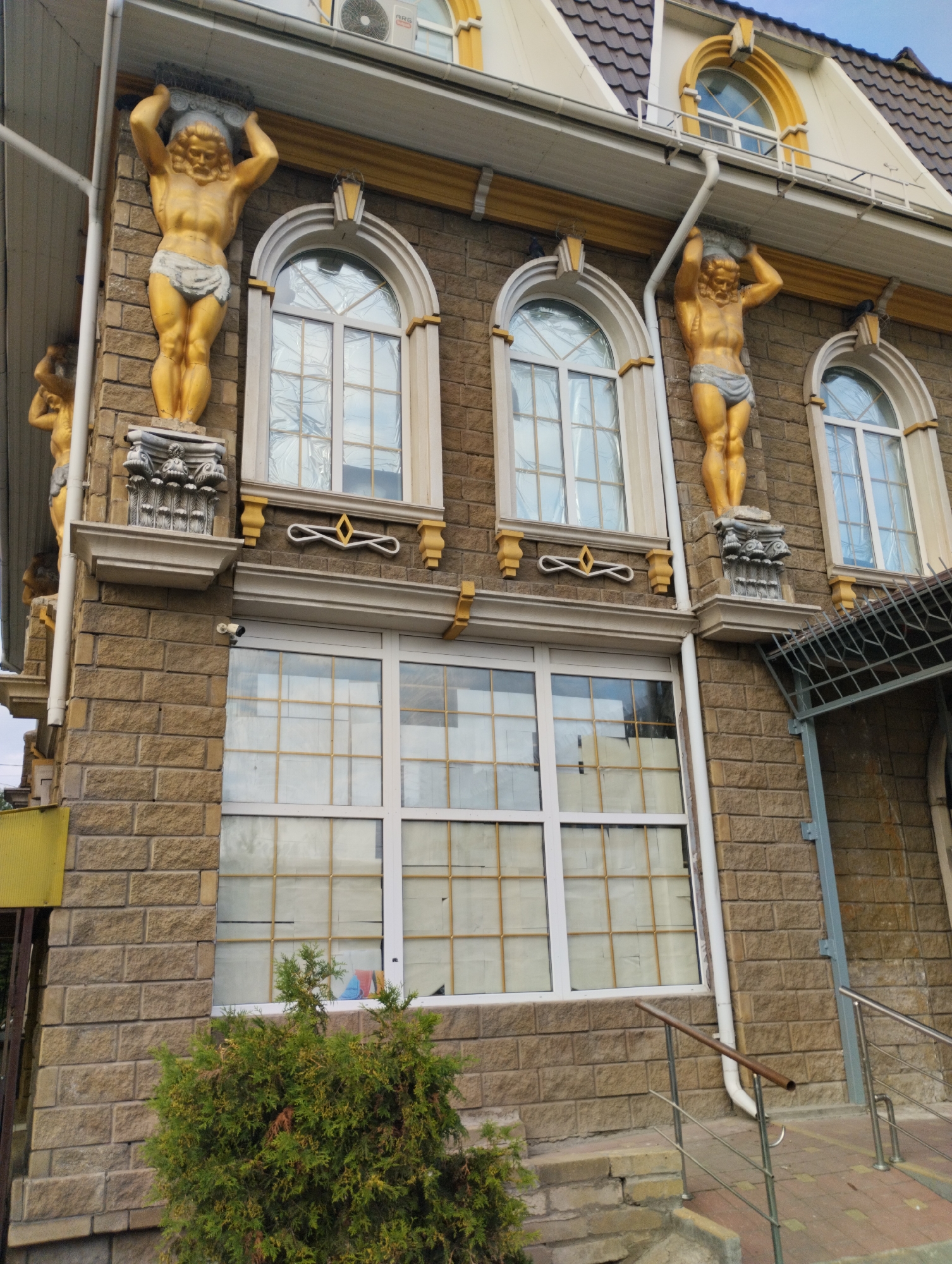 A golden statue of Atlas, a muscular male figure with a beard and loincloth, supports a decorative element on the facade of a building in Kostanay, Kazakhstan. The statue is positioned on a pedestal with ornate carvings, and there is another similar statue visible to the left, partially obscured. The building's facade is constructed from light brown brickwork and features arched windows with white frames. A security camera is visible mounted on the brick wall below one of the windows. In the foreground, a green evergreen shrub is present, and a staircase with metal railings leads up to the building entrance. The sky above is a pale blue.