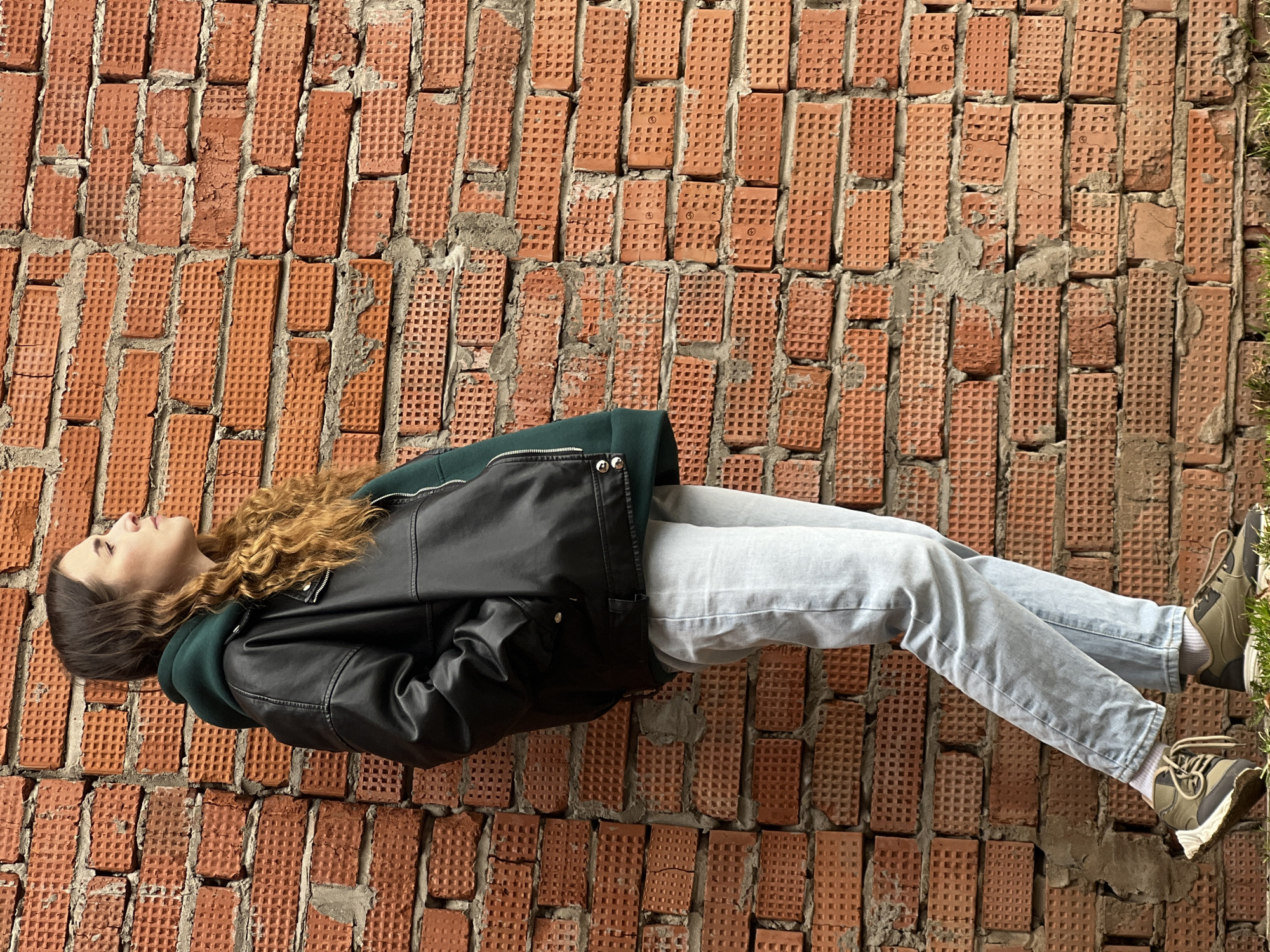 A young woman is reclining against a brick wall. She is wearing light blue jeans, and dark green and black sneakers. She is also wearing a dark green hoodie underneath a black, glossy jacket. She has long, curly, brown hair. Some green vegetation is visible at the far right edge of the wall. The wall appears to be constructed of textured orange-red bricks. The location is in Votkinsk, Russia.