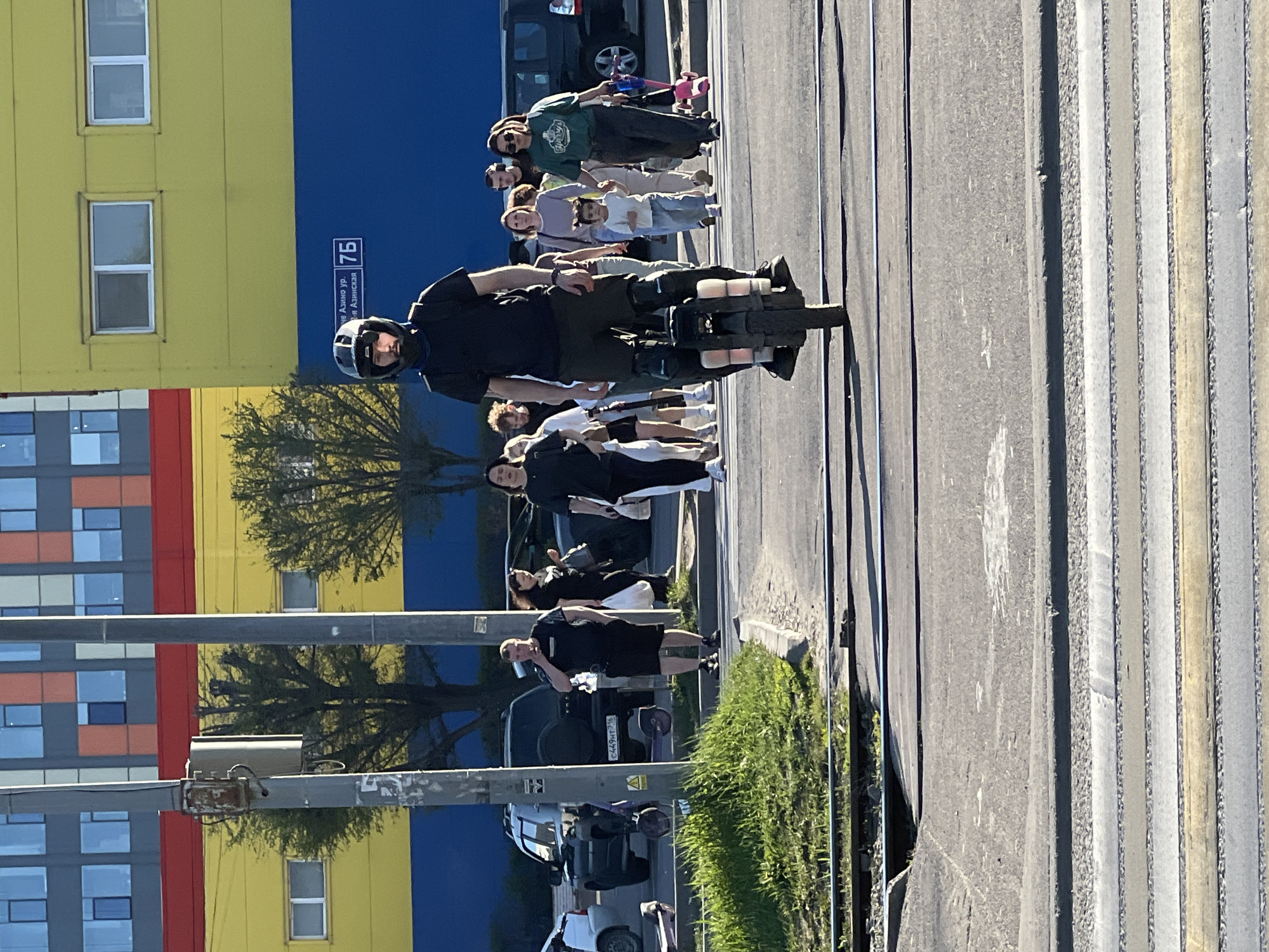 The image shows a street scene in Kazan, Russia. A man wearing a helmet and dark clothing is riding a motorized unicycle on a pedestrian crossing. Several people are crossing the street along with him, including families with children and individuals in casual attire. The background features a multi-colored building in shades of blue, yellow, red and orange. A sign is visible on the side of the building with the address "ул. Азино, ул. Азинская 7Б". Parked cars and a patch of grass are visible along the side of the road. The street is well-lit, suggesting it is daytime.