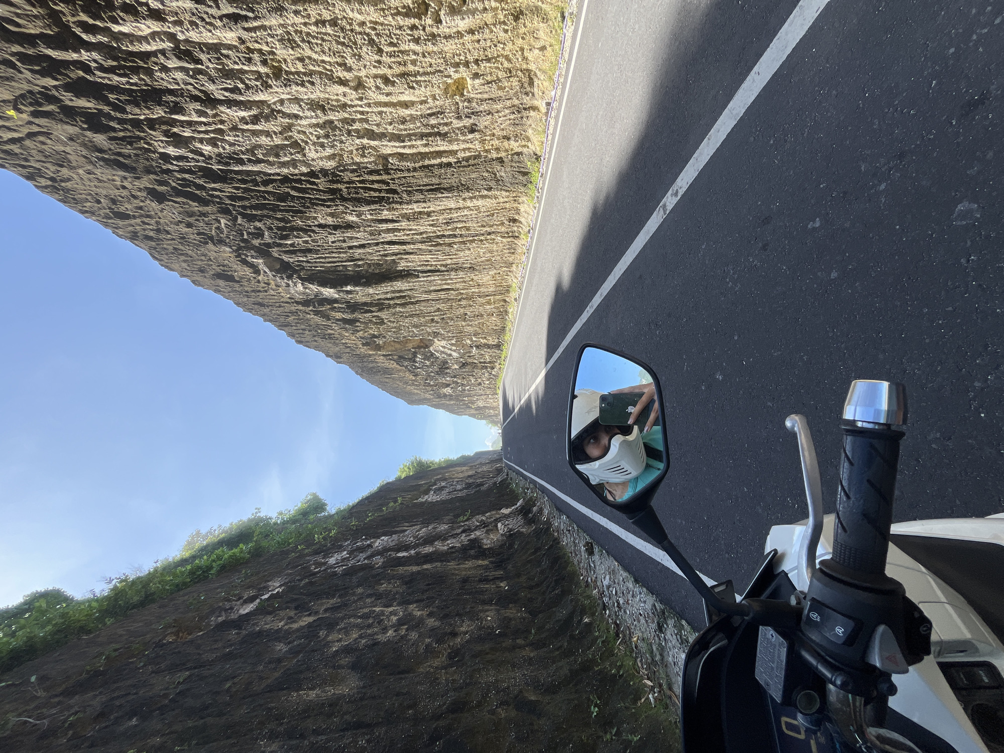 The photograph depicts a view from a moving motorcycle on a paved road in Kuta Selatan, Indonesia. High rock walls flank the road on both sides, with exposed geological stratification evident on the wall to the left and vegetation clinging to the wall on the right. The sky is visible above the cliffs. A white line marks the edge of the road. The motorcycle's right side mirror reflects the rider, who wears a mask and helmet and holds a smartphone. The motorcycle's handlebars, grip, brake lever, and some instrumentation are visible.