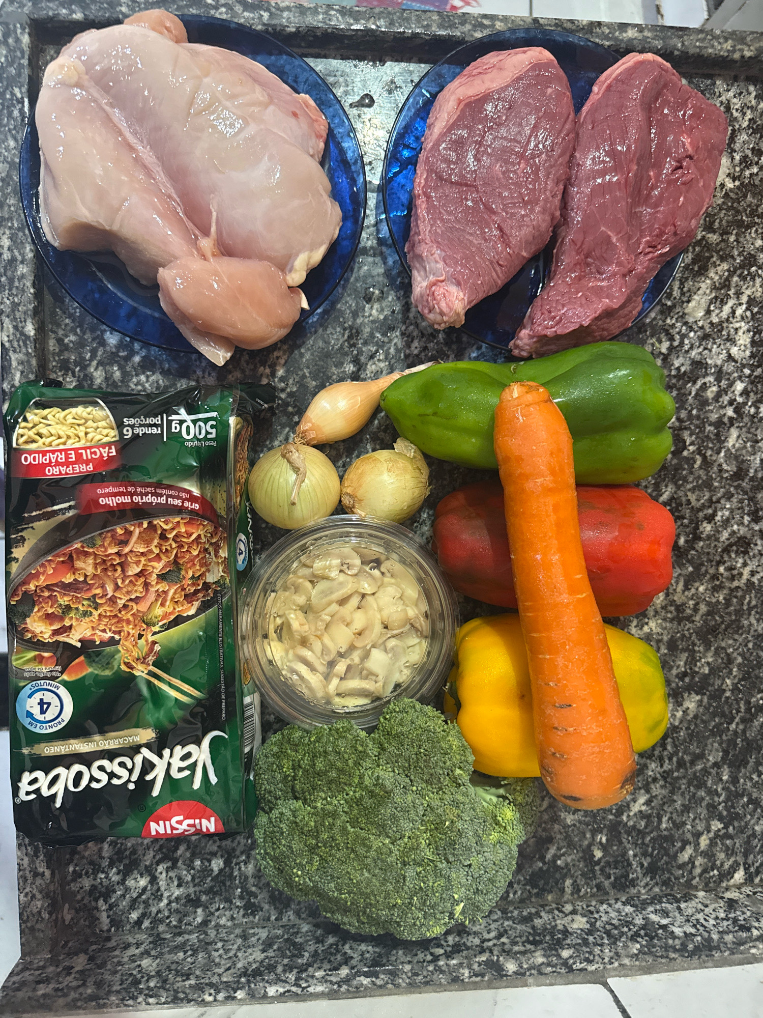 The image depicts a collection of raw ingredients laid out on a speckled gray countertop. In the upper left, a halved raw chicken is presented on a blue plate. To its right, on another blue plate, are two cuts of raw red meat. Below the chicken and meat, a package of Nissin Yakissoba noodles is visible. To the right of the noodle package, various vegetables are arranged: a bunch of broccoli, a large carrot, a green bell pepper, a red bell pepper, and a yellow bell pepper. Two yellow onions and a shallot are positioned near the top center. A clear plastic container filled with sliced mushrooms is also present. The lighting suggests an indoor setting, possibly a kitchen. There are no people or actions depicted in the image. The countertops appear to be made of granite or a similar composite material. The image does not provide any information or context about the city of Nilópolis, Brazil, or any specific location within it.