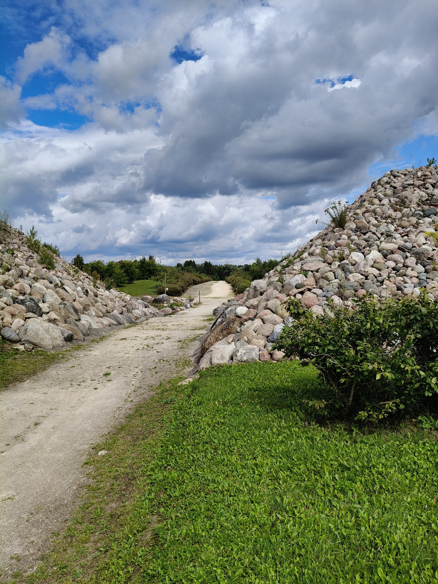 The image shows a dirt path winding through a landscape characterized by large piles of rocks and lush green grass under a cloudy sky. On either side of the path are imposing mounds of rounded stones, some small and others quite large, appearing to form artificial hills. The path itself is a light brown, dusty color, with patches of sparse vegetation growing along its edges. To the right of the path, a dense green bush is visible. In the distance, trees form a natural backdrop, and the sky is filled with a mix of dark, heavy clouds and lighter, broken patches revealing blue sky. The scene appears to be an outdoor recreational area or park.