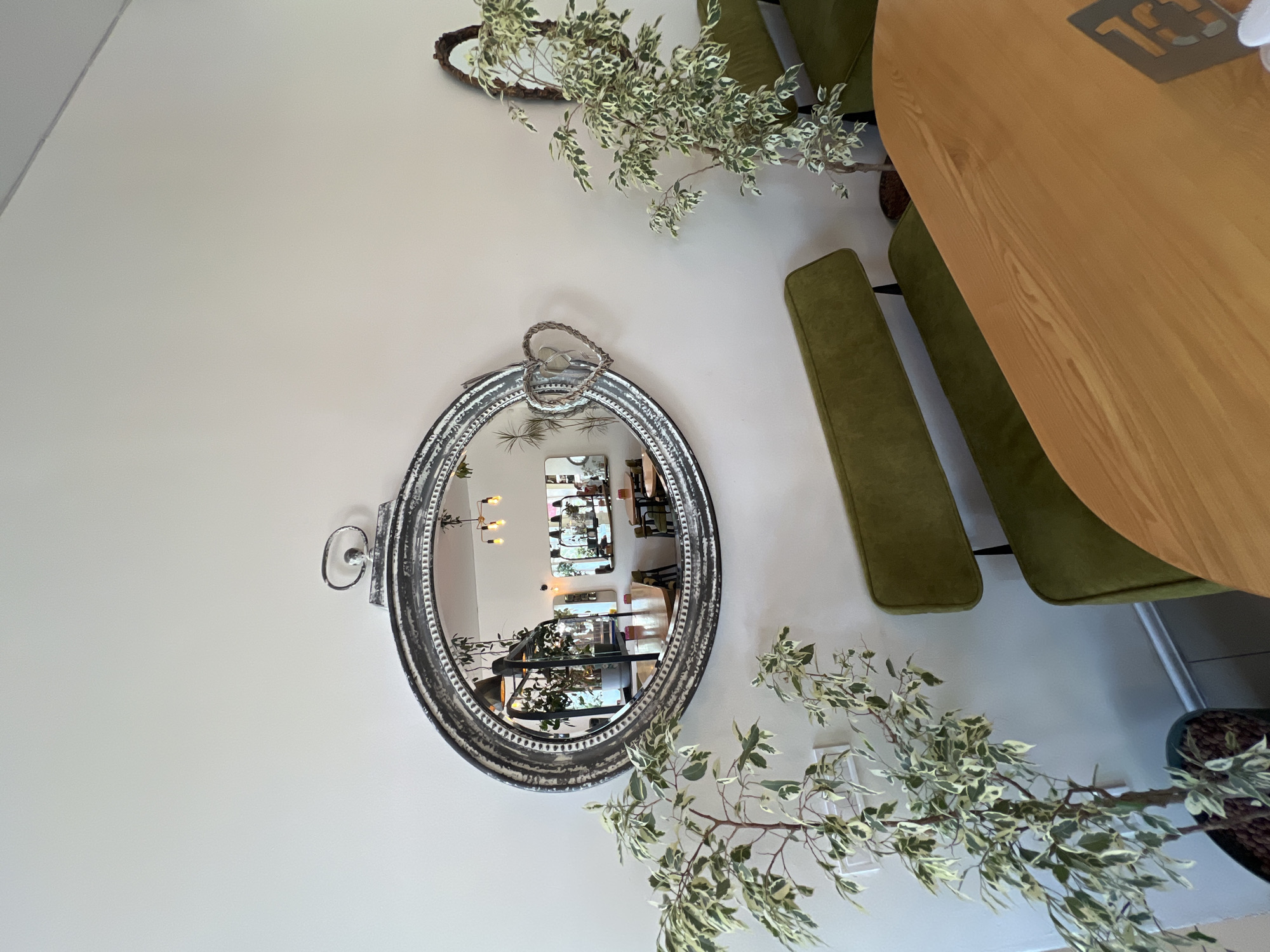 The image depicts the interior of a cafe in Odesa, Ukraine. A large, oval mirror with an ornate, distressed silver frame hangs on a light-colored wall. A decorative heart made of intertwined twigs is attached to the lower right side of the mirror. The mirror reflects the interior of the cafe, showing tables, chairs, and more plants. In the foreground, two potted plants with variegated leaves are positioned to the left of the mirror. A green upholstered bench seat is visible below the mirror and a portion of a light-colored wooden table occupies the lower right side of the frame. A smaller, oval mirror with a dark, rustic frame is visible on the wall to the right of the large mirror.