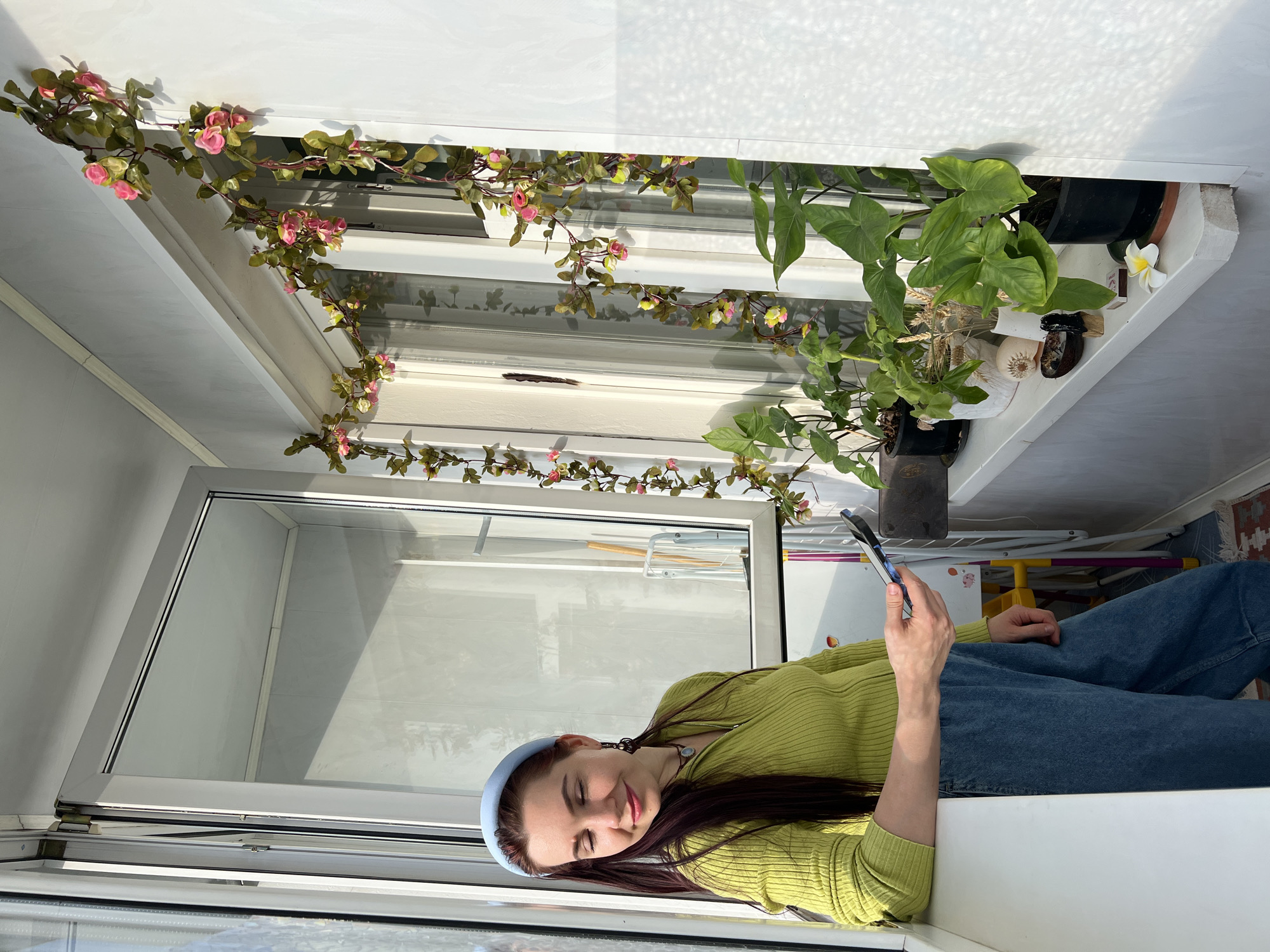 A woman is sitting in an indoor space with a large window. She is wearing a light green sweater, blue jeans, and a light blue headband. She is looking at a cell phone in her hand. The window is decorated with a vine of pink flowers and potted plants. The city and location are unknown.