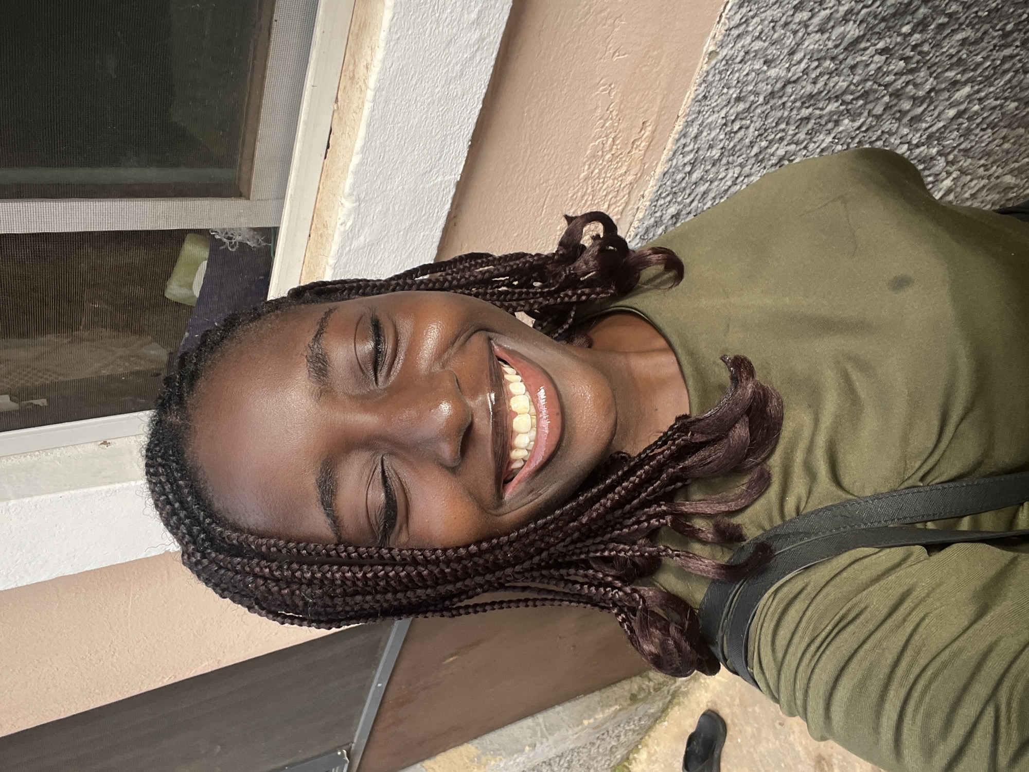 The image is a vertical, close-up portrait of a young Black woman captured outdoors in bright daylight. She is depicted from the chest up, with her head slightly tilted and a radiant, wide smile that causes her eyes to close, suggesting genuine laughter or joy. Her dark hair is styled in numerous small box braids, which frame her face and extend over her shoulders.
She is wearing a simple, olive green or khaki-colored short-sleeved top, with a black strap, likely from a shoulder bag, visible across her right shoulder. The bright natural light illuminates her face, highlighting her smile.
The background features various architectural elements. To the left, a window with a screen mesh is visible. Behind her, sections of an exterior building wall display different textures and colors: a light beige or off-white painted area, a peach-colored segment, and a prominent grey, textured surface, possibly pebble-dashed or rough concrete, on the upper right. A dark, horizontal ledge is visible beneath the window. Part of a black slipper or shoe can be seen in the bottom right corner of the frame.
The image conveys a strong sense of happiness and warmth, emphasized by the woman's joyful expression and the sunny conditions. The bright sunlight indicates a clear daytime setting. The background elements suggest a casual outdoor environment, consistent with a building exterior in Abuja, Nigeria, as provided by the location context. There is no visible text in the image.