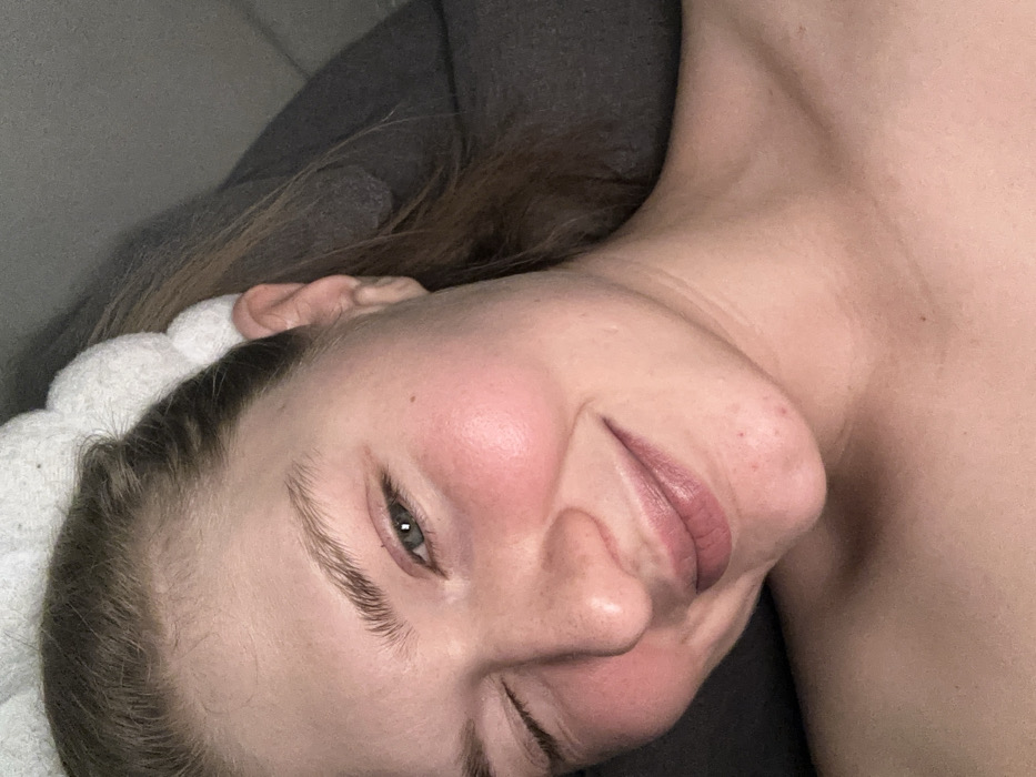 A fair-skinned person wearing a white headband smiles slightly, displaying flushed cheeks and a relaxed expression. Their gaze is directed towards the viewer with one eye partially closed. The background consists of gray, possibly indicating a pillow or cushioned surface. The individual's head is tilted to the right, revealing part of their shoulder and collarbone.