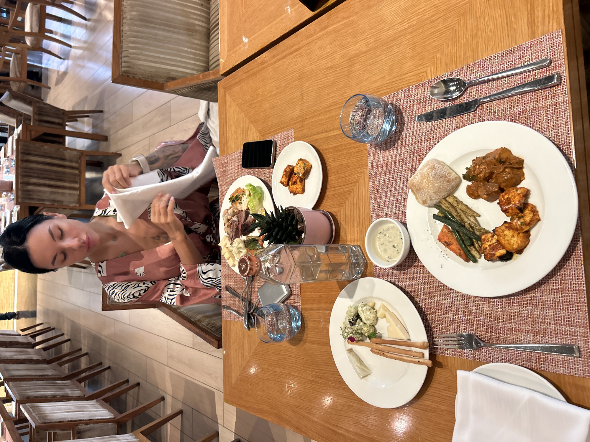 The image shows a woman sitting at a table laden with food at what appears to be a restaurant in Pune, India. The woman is wearing a patterned robe and is in the process of folding a white napkin. On the table, there are several plates of food, including what looks like a cheese platter, grilled vegetables, and some kind of curry. Drinks, cutlery, and a mobile phone are also present. The restaurant's interior is visible in the background, with rows of chairs and tables suggesting a spacious dining area. The overall impression is of a meal being enjoyed in a well-appointed establishment.