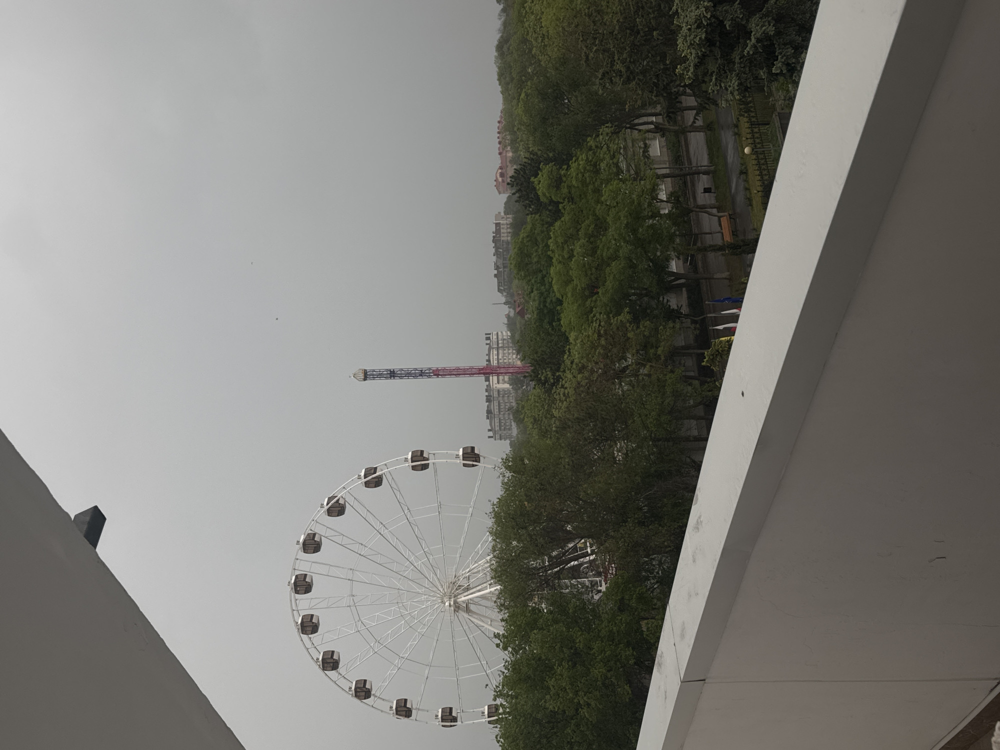The image shows a partially obscured view of the city of Yevpatoriya, Ukraine. A large Ferris wheel with enclosed passenger cars is visible on the left, partially obscured by trees. In the background, a tall, vertical amusement park ride stands between buildings and trees. The sky is overcast. In the foreground, portions of a white, modern-looking building or structure frame the view on both sides of the image.