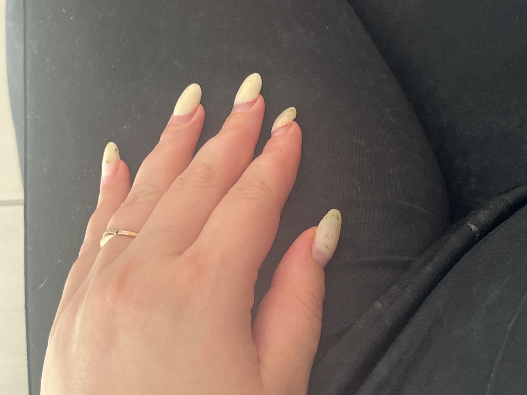 The image shows a close-up view of a person's left hand with light-colored nails, resting on a dark fabric surface, possibly clothing. A thin gold ring with small stones is visible on the ring finger. The nails appear to have a light yellow polish with some dark specks on them, and there are signs of wear and tear, particularly on the thumb nail which is chipped. The background is out of focus and appears to be a solid dark color. No specific location in Chelyabinsk, Russia, can be identified from the image.