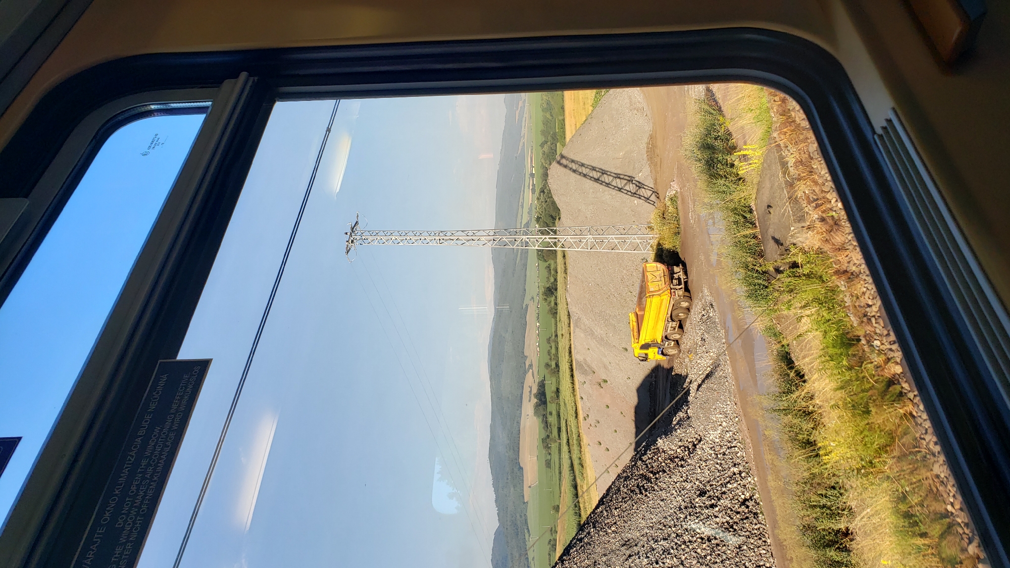 This is a photograph taken from inside a train compartment, looking out of the window at a rural industrial landscape. The scene features a bright yellow dump truck parked on a dirt road next to large piles of gravel. A tall, metallic power line pylon stands prominently in the middle ground. In the background, rolling green hills and fields stretch towards a clear blue sky. The window itself has some reflective text, likely a warning about not opening the window due to air conditioning. The overall impression is one of a sunny day in an industrial or construction area located in a rural setting. The specific location of Spišský Štiavnik, Slovakia, is relevant context for this scene, suggesting a geographical setting within that region.
