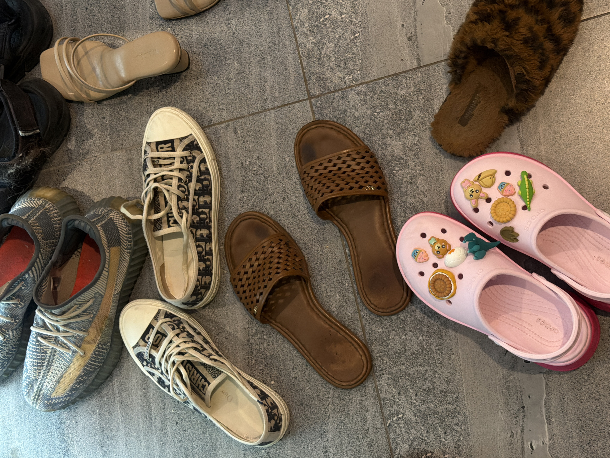 The image depicts an assortment of footwear arranged on a gray tiled floor, likely within an indoor space. The footwear includes: two pairs of sneakers with blue and beige patterned exteriors and white laces; a pair of gray and beige Adidas Yeezy sneakers; a pair of tan leather sandals; two brown woven leather sandals; a pair of brown faux-fur lined slippers; and a pair of pink Crocs adorned with Jibbitz charms. The location is not discernable as Kerobokan, Indonesia, based solely on the visual information provided. There are no individuals visible in the scene, and the only activity implied is the placement of the shoes on the floor.