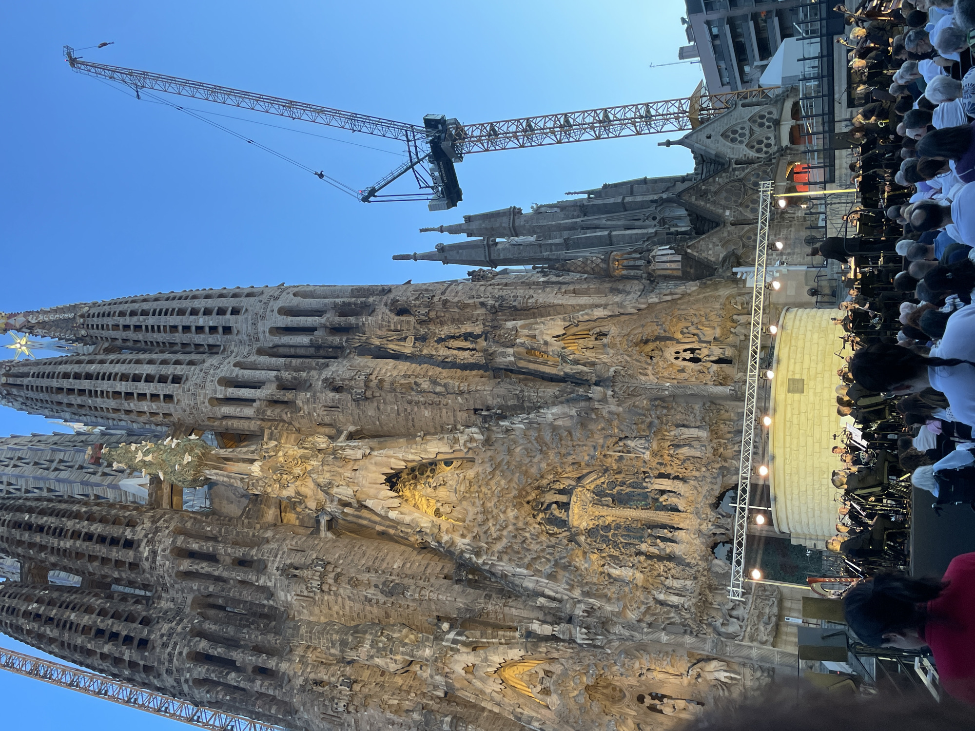 The image depicts a wide shot of the Sagrada Familia, a large Roman Catholic basilica in Barcelona, Spain, with a concert performance taking place in front of it. The basilica's intricate facade, characterized by detailed sculptures and spires, is prominently featured. A tall construction crane is visible to the right of the basilica, suggesting ongoing construction or renovation work. In the foreground, an orchestra is seated on a stage, with musicians playing instruments. A conductor stands at the front, directing the ensemble. A crowd of people are seated and standing, watching the performance, with some in wheelchairs. The scene is illuminated by stage lights and the ambient light of early evening, with a clear blue sky overhead. The overall atmosphere appears to be a cultural event held at a historic landmark.