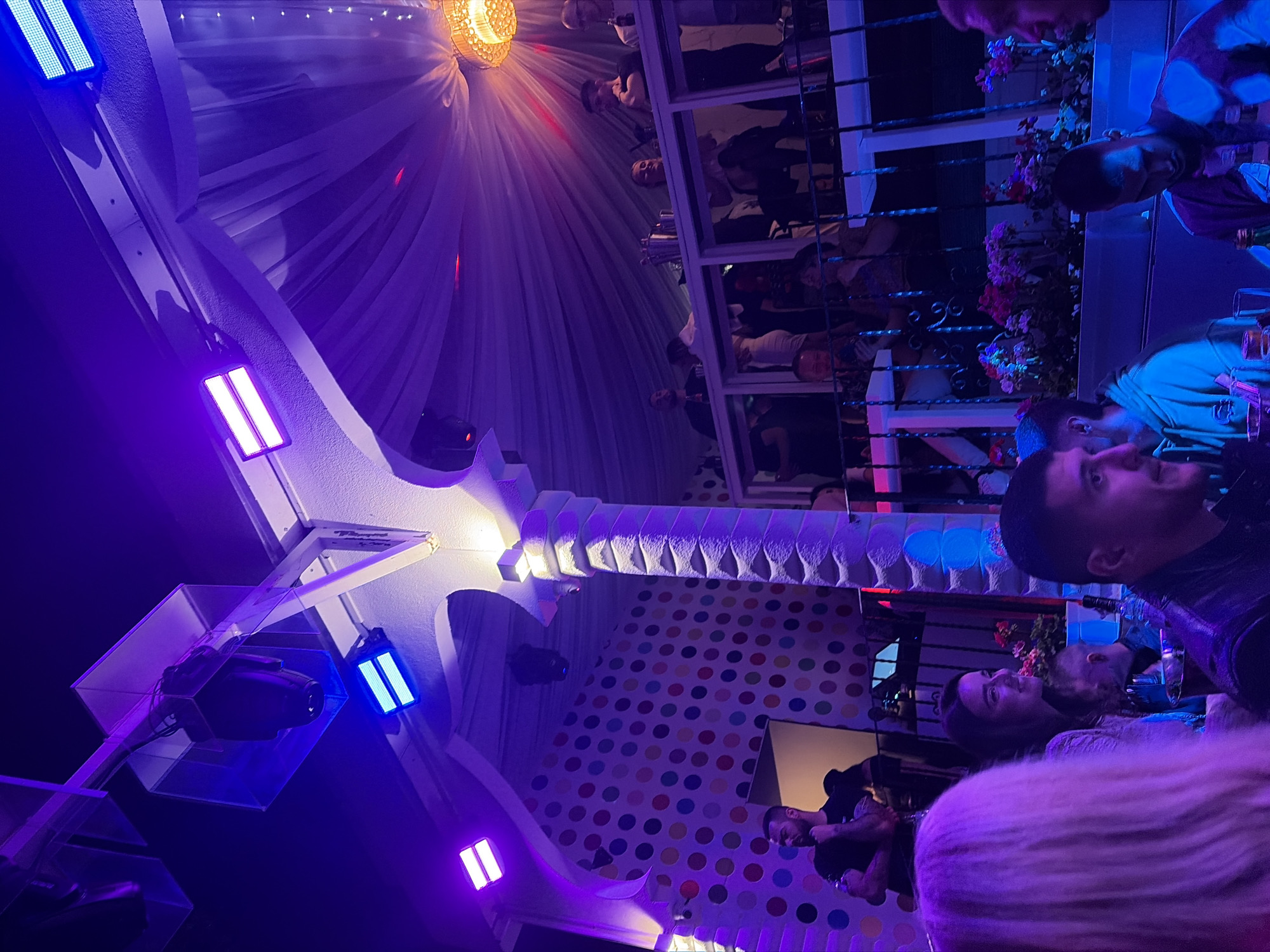 This vertically oriented image captures a lively, brightly lit nighttime scene inside an entertainment venue, possibly a nightclub or lounge in Varna, Bulgaria. The ambiance is dominated by vibrant purple and blue artificial lighting from LED panels mounted on structural elements. A warm-toned, ornate chandelier hangs from a draped white fabric canopy on the ceiling, adding another light source.
Numerous people, primarily young adults, are present, socializing and enjoying the atmosphere. Some are seated at tables on a lower level, while others are visible on an upper level or balcony, leaning against a white railing adorned with potted plants bearing red and pink flowers. Architectural details include white beams and columns, one featuring a distinct wave-like pattern. A large wall in the background is decorated with a colorful polka-dot pattern in red, yellow, blue, green, and purple. A projector is mounted on a ceiling support.
The overall impression is one of a social gathering in a modern, stylish venue after dark. No visible text is present in the image.