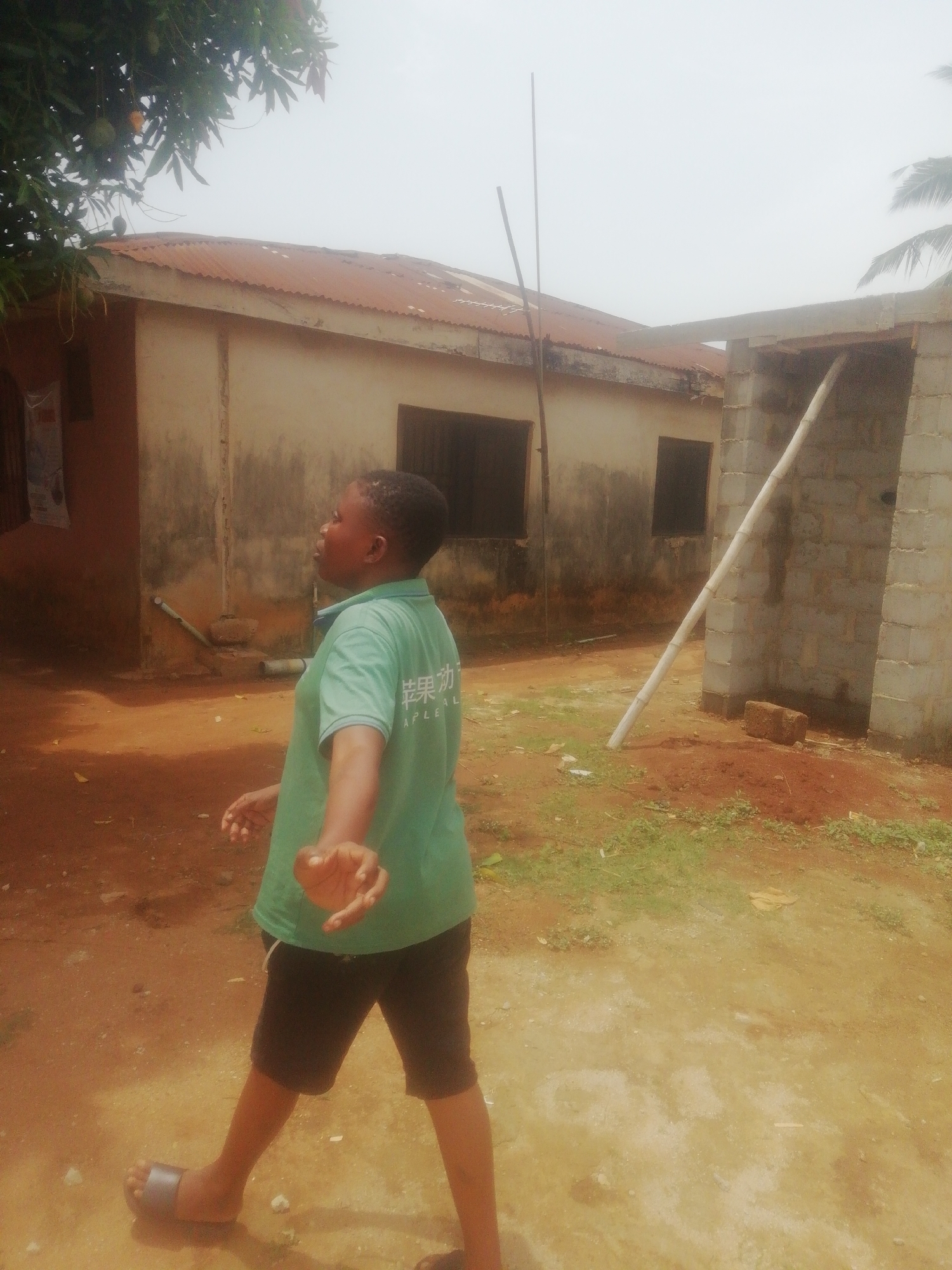 A full outdoor shot shows a child in the right foreground walking away from the camera with their left hand stretched out. They wear a bright green t-shirt with what appears to be writing in another language on the back, along with black shorts and dark-colored sandals on their feet. To the right, the unfinished concrete structure of a building under construction is visible. Next to the child is a path covered in reddish-brown dirt with patches of short green grass. In the background is a one-story building with dirty beige-colored walls and a rusty red roof. Two bare poles extend from the roof. The sky is mostly white with only the tops of a few trees barely visible. The image is taken outdoors in the city of Unknown, Unknown.