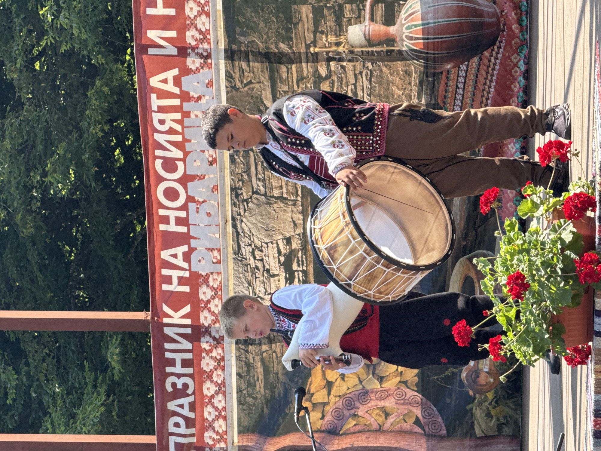 Two young boys are performing on a wooden stage in Ribaritsa, Bulgaria, during a festival. The boy on the left, facing right, is playing bagpipes. He wears a traditional white shirt with embroidered sleeves and a red vest. The boy on the right, facing left, is holding a large drum. He is dressed in a similar traditional outfit, including a white shirt, a black vest with colorful trim, and brown pants. Behind them, a banner with white lettering on a red background reads "ПРАЗНИК НА НОСИЯТА И Н." (Festival of the Carriers and the New Year/Tradition). The background depicts a stone wall and a stage decoration featuring traditional patterns and a large, ornate vase. In the foreground, potted red geraniums add a splash of color to the scene. The event appears to be taking place outdoors on a sunny day.