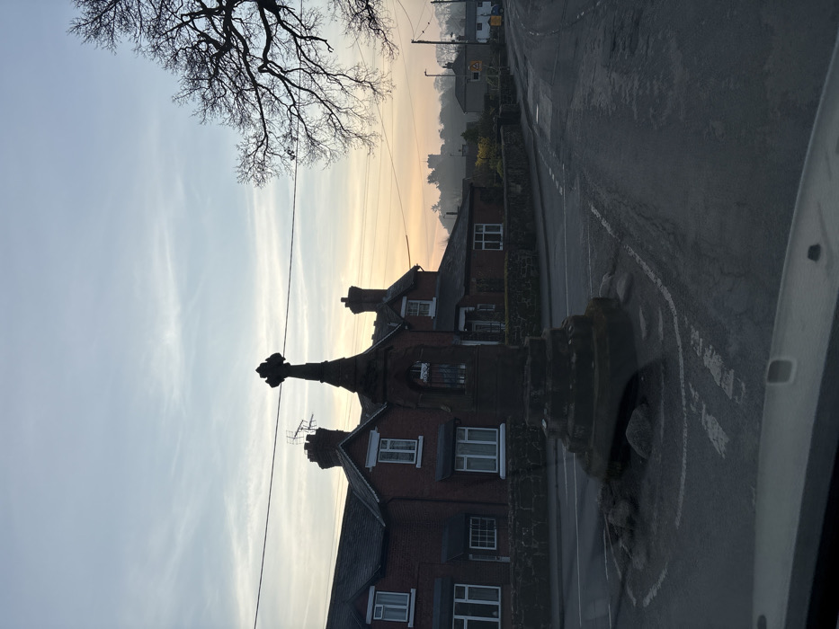 The image depicts a road junction in Ruyton-XI-Towns, United Kingdom, at either dawn or dusk. In the immediate foreground, a dark, weathered stone market cross stands on a small, circular, rock-lined traffic island. The cross features a tiered octagonal base, a central shaft with an arched niche containing vertical bars on one face, and a finial at its apex.
Behind the monument, to the left, are several two-story red-brick buildings with dark-tiled roofs and multiple white-framed sash windows. An antenna is visible on a chimney stack of the leftmost building. Overhead, multiple horizontal utility lines traverse the upper portion of the frame. To the right of the cross, a large deciduous tree, currently bare of leaves, presents a dark, intricate silhouette against the sky.
The asphalt road, which appears damp or reflective, encircles the monument and extends into the background, displaying faded white road markings. In the distant background, a large stone structure, possibly a church or castle, is visible atop a hill, partially obscured by atmospheric haze. The sky transitions from a pale blue overhead to a soft orange and yellow hue near the horizon. The vantage point is from inside a vehicle, with a portion of the dashboard visible along the bottom edge of the image. No human figures or active vehicles are present in the scene.