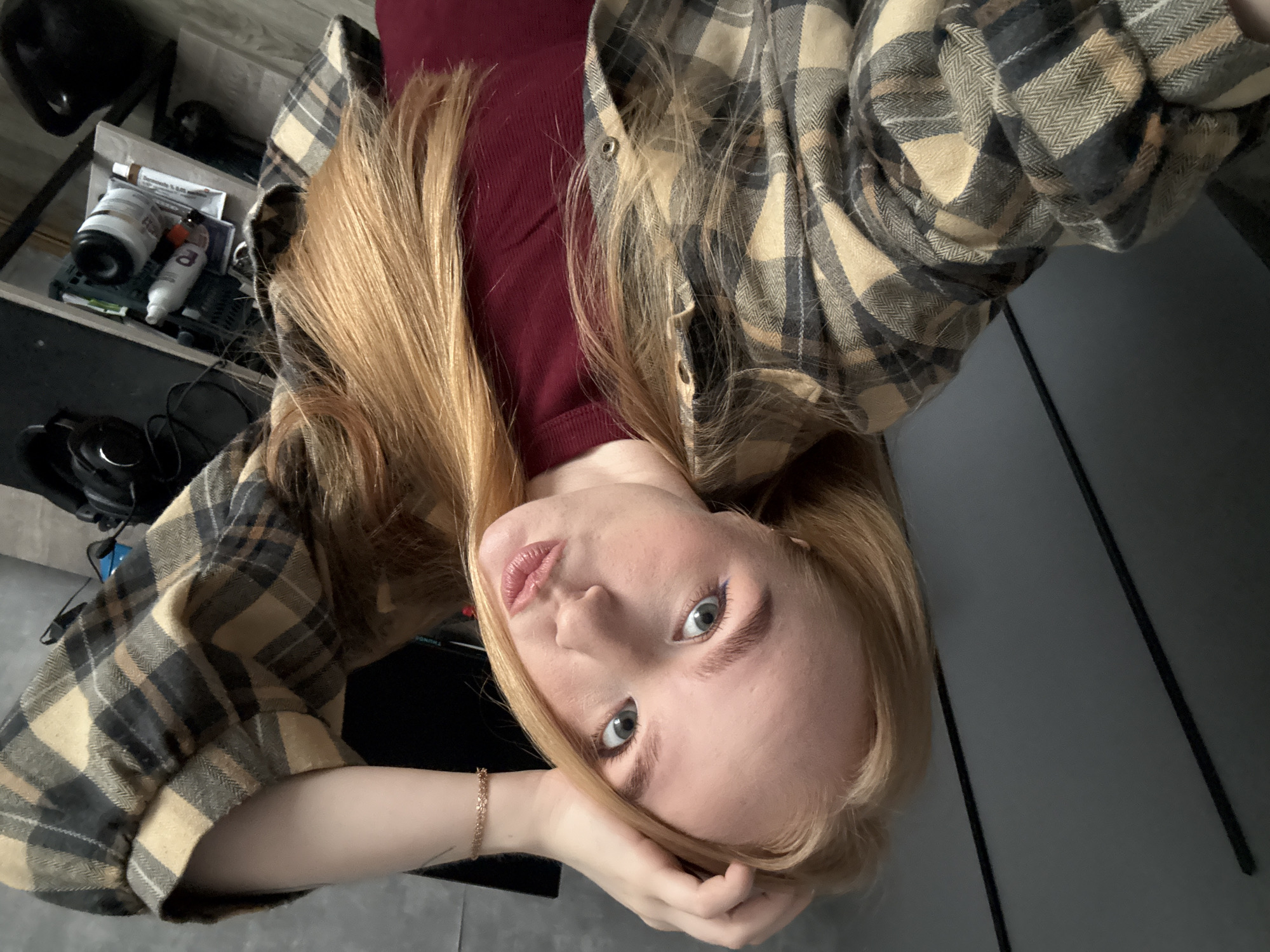 A young woman is shown in a close-up, angled shot, looking up at the camera. She has long, light blonde hair, fair skin, and blue-grey eyes. She is wearing a maroon, possibly corduroy, top and a light brown and dark grey plaid flannel shirt. Her left arm is visible, resting near her face, with a gold bracelet on her wrist. The background reveals a cluttered space with a black shelf containing various items including bottles, packaging, and headphones. A grey surface is also visible, which appears to be a floor. The location and city of Chaikovsky, Russia, cannot be verified from this image.