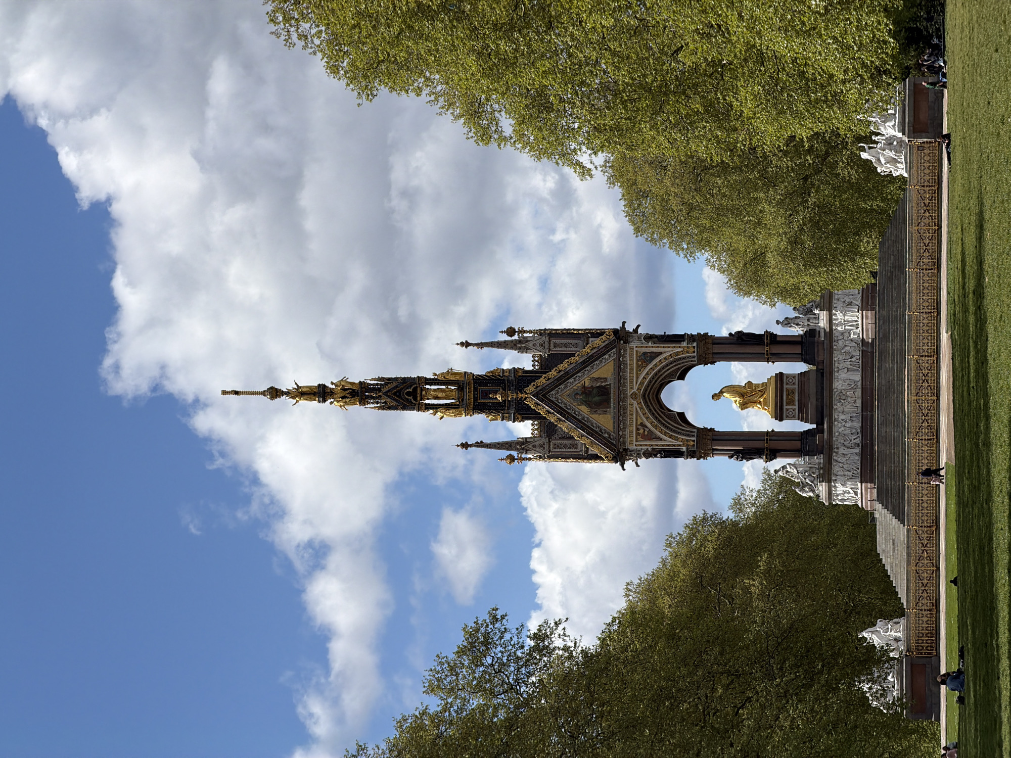 The image captures the majestic Albert Memorial, a large Gothic Revival monument, centered within a spacious park setting. The memorial features intricate dark spires and arches adorned with numerous golden accents, including a prominent golden statue of Prince Albert seated beneath one of its central arches. White marble sculptures are arranged around the base of the monument, adding to its grandeur.
Lush green trees with vibrant spring foliage frame the upper corners and line the sides of the scene, suggesting a natural park environment. On the right side, a wide expanse of bright green grass stretches out, with a slight upward slope towards the memorial's base.
The sky above is a mix of clear blue patches and fluffy white clouds, indicating a bright, pleasant day, likely late morning or afternoon given the consistent lighting. Several small figures of people are visible on the grass and near the monument's steps; some are seated on the lawn, seemingly enjoying the peaceful atmosphere. The overall scene conveys a serene and leisurely mood in a significant public space in London, United Kingdom. No discernible text is visible.
