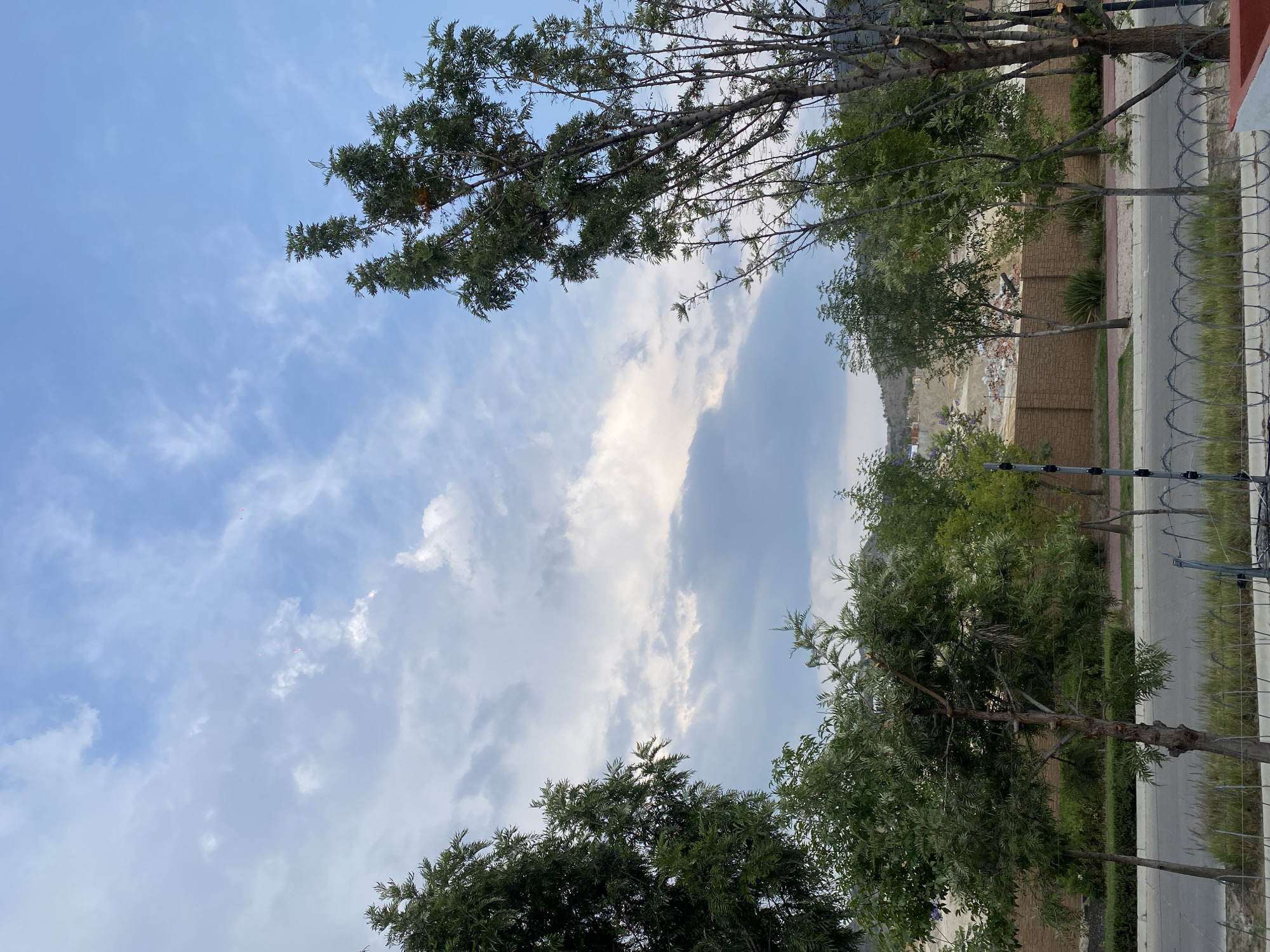 The image shows a street in an urban environment, possibly on the outskirts of a city. The view is from ground level, looking upward. The sky is blue with scattered white clouds. In the foreground and partially obscuring the sky, are the leafy branches of trees. To the right, a brick wall stands alongside a road. A fence with barbed wire sits on the right. Beyond the wall, there's a disturbed landscape, possibly a construction site or a dump. No people are visible in the image.