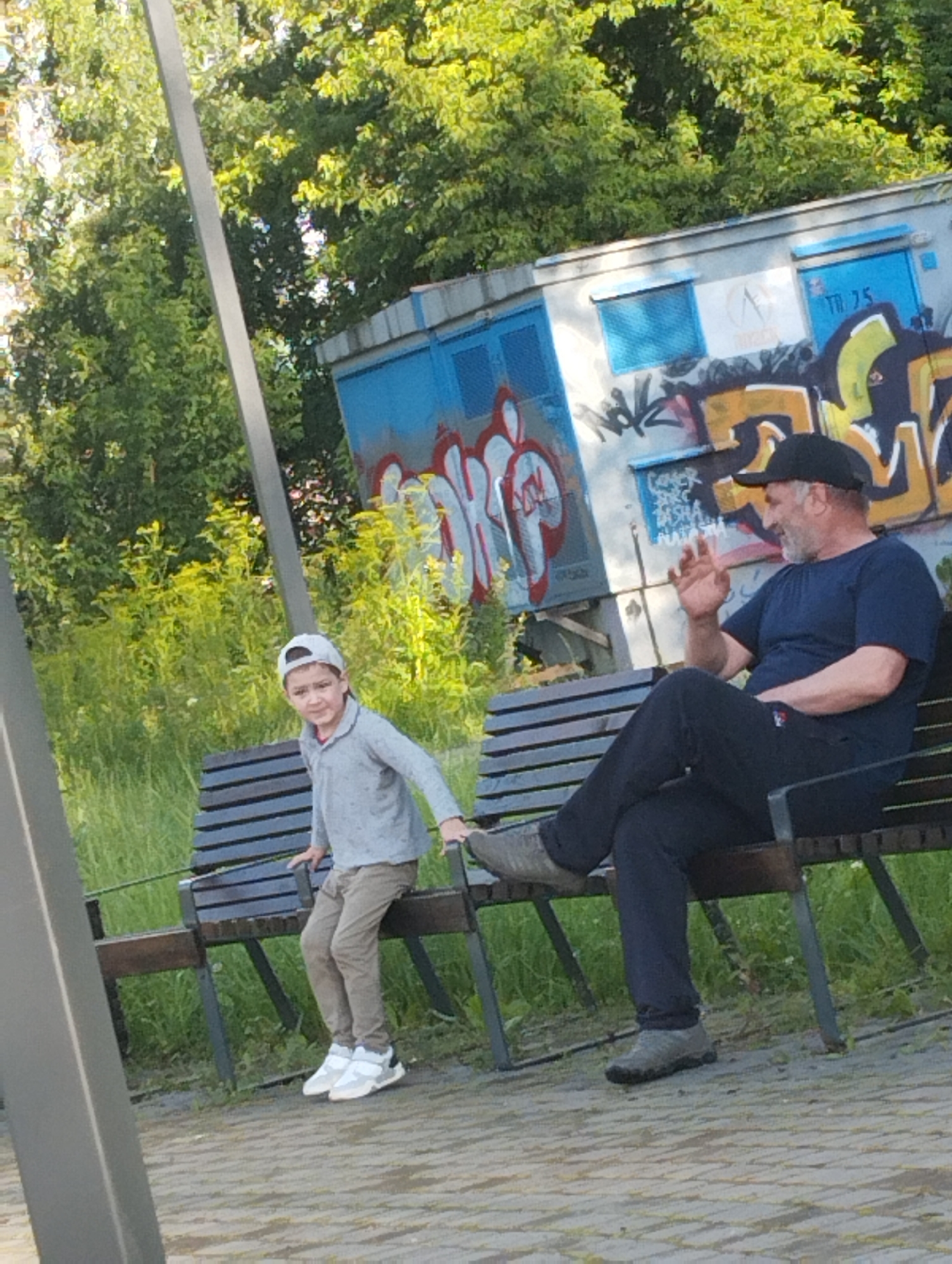 In Kingisepp, Russia, on a sunny day, a man and a boy are seated on a park bench. The man, wearing a dark blue t-shirt and a black baseball cap, is on the right side of the bench. His left leg is crossed over his right, and he is gesturing with his right hand. The boy, on the left side of the bench, is wearing a light grey long-sleeved shirt, tan pants, a white baseball cap, and white and grey sneakers. He is looking towards the camera. Behind them, a blue structure with graffiti is visible, along with lush green trees and foliage. The ground is paved with light-colored bricks.