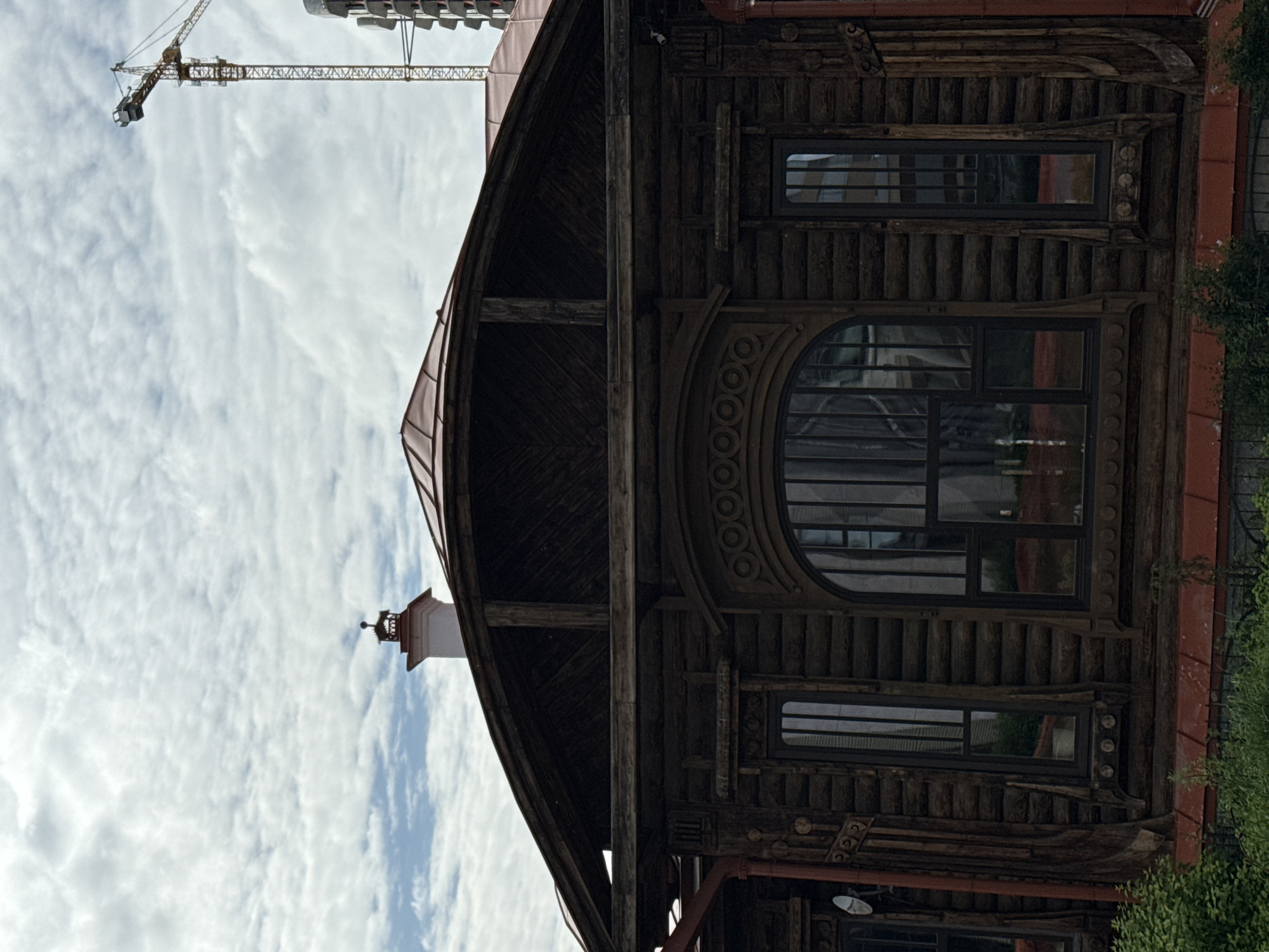 The media file displays an exterior view of a historic wooden building with ornate carvings and multiple windows. The building has a curved, dark wooden facade and a reddish roof. A large arched window with intricate detailing is a prominent feature, alongside several smaller rectangular windows. The architecture suggests a traditional style, possibly with a historical significance.
To the left of the building, a construction crane is visible against a cloudy sky. The sky is filled with white, wispy clouds, indicating daytime. In the distance, parts of a modern building are also visible, suggesting the juxtaposition of old and new architecture. A white chimney with a decorative top extends from the roofline of the wooden building. Green foliage is present along the right edge of the frame, indicating an outdoor setting with some vegetation.
The image appears to be taken in Yekaterinburg, Russia, as stated in the location context. The overall mood is one of historical preservation amidst ongoing development. No people or specific activities are depicted; the focus is on the architectural details of the building and the surrounding environment. There is no visible text in the image.