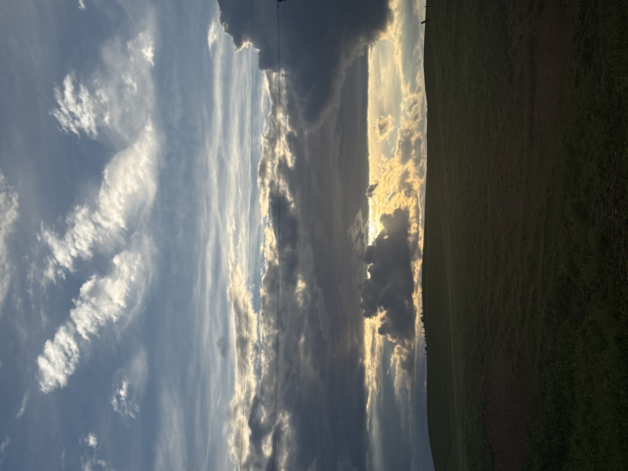 The image shows a field of green grass taking up the right side of the frame. To the left of the field, a cloudy sky is visible. The clouds range in color from white to dark gray. Sunlight peeks through the clouds in the center of the frame, creating a bright spot. Power lines stretch across the sky near the center. The image is rotated 90 degrees clockwise. There is no evidence to suggest this image was taken in Batmany, Russia.