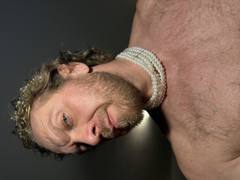 A close-up portrait depicts an adult male from the chest up, with light brown, wavy hair and a full beard. He wears a gold-colored tiara with clear stone embellishments on his head and a multi-strand white pearl necklace tightly around his bare neck. A small, square, silver-colored stud is visible in his right earlobe. His chest and right shoulder are exposed, revealing body hair. The man has blue eyes and a stern expression, with furrowed brows and a slightly downturned mouth, directing his gaze towards the viewer. The background is a plain, dark gray or black, suggesting an indoor or studio environment. A strong light source illuminates the left side of his face and body, creating shadows on the right. The specific location within the city of Saint Petersburg, Russia, cannot be determined from the visual content of the image.