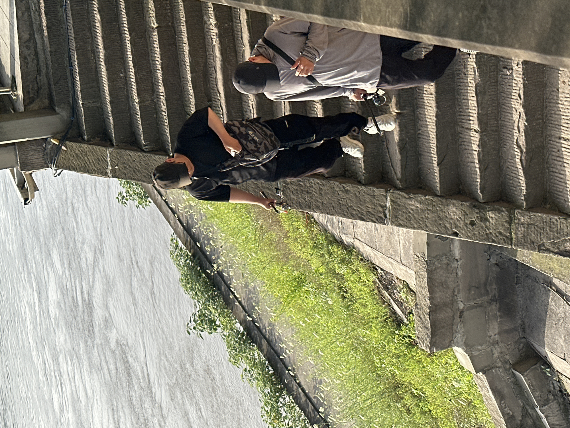 Two people are fishing from a high stone platform or bridge. One person is wearing a black outfit and the other is wearing gray, both with dark caps and white shoes. The platform overlooks a wall covered in green vegetation and a large body of water. The bridge or platform is constructed of gray stone, and the city of Jiangjin, China, is mentioned as the location.