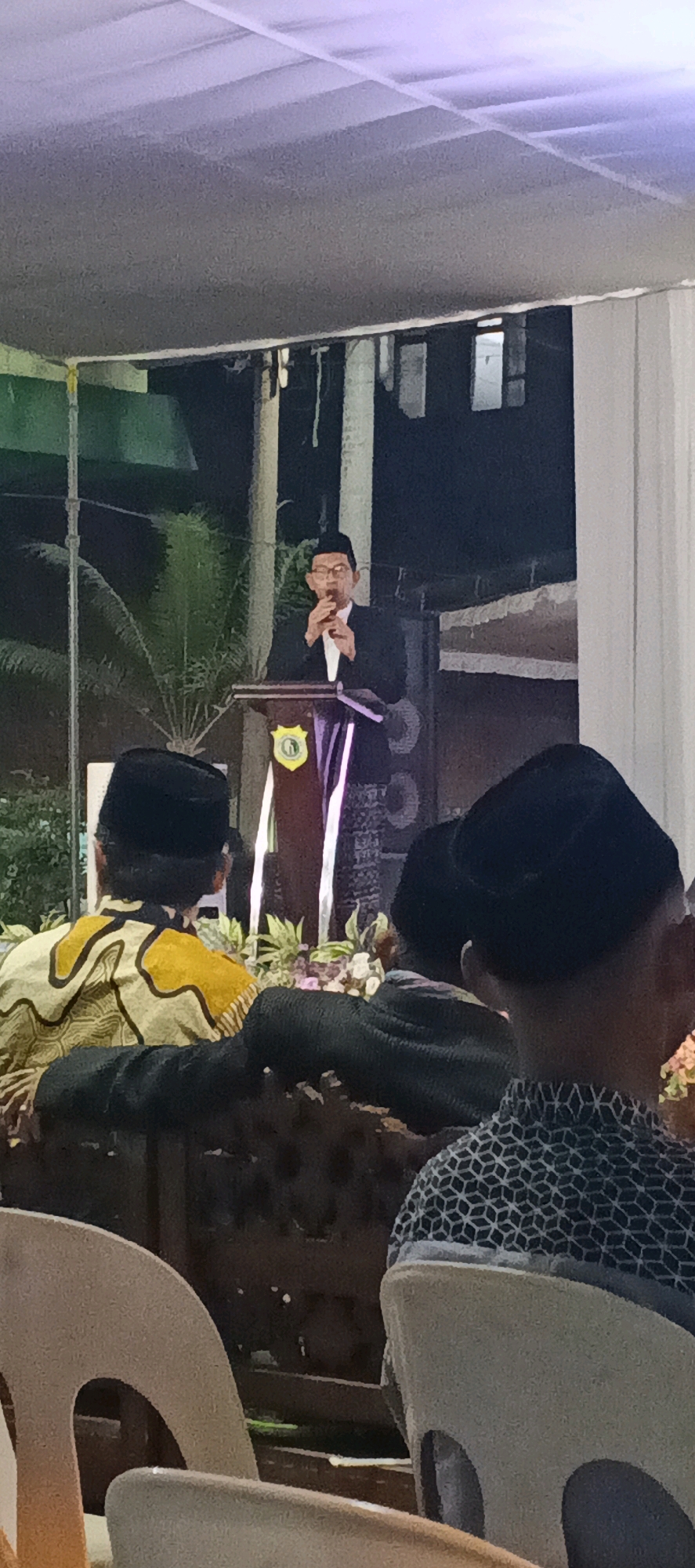 The image depicts an event taking place outdoors, likely in the evening, at a location in Sungelebak, Indonesia. A man wearing a black cap, glasses, and a black suit over a white shirt is speaking at a podium. The podium is decorated with a crest and purple fabric. Behind the speaker, there are palm trees and some building structures. In the foreground, several people are seated, facing away from the camera. One person in the front row wears a yellow and black patterned shirt and a black cap. Another person in the foreground is partially visible, wearing a patterned shirt and a black cap. The seating appears to be made of chairs and possibly benches. The overall atmosphere suggests a formal gathering or ceremony.