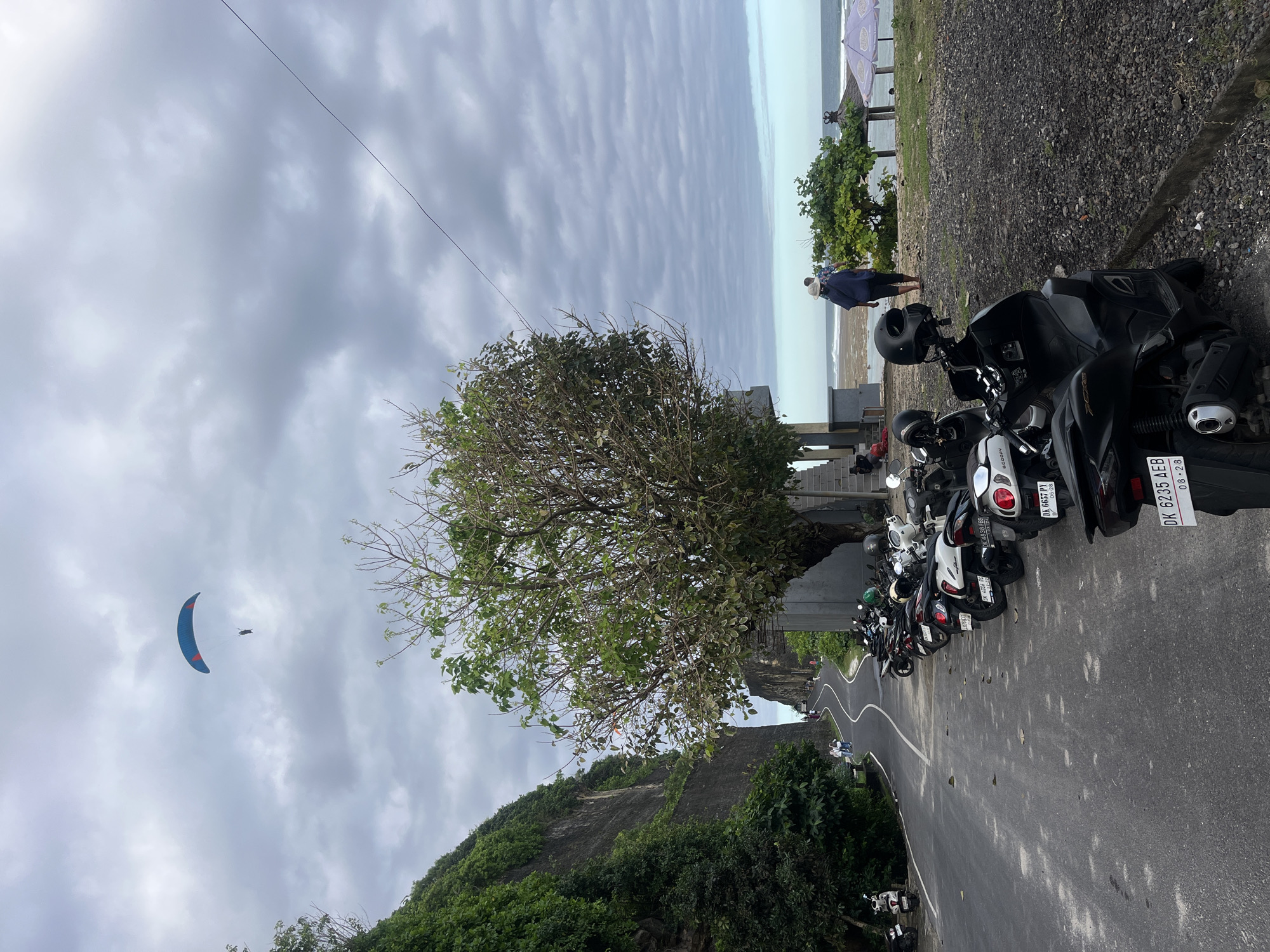 The image depicts a coastal scene in Kuta Selatan, Indonesia. A winding asphalt road occupies the lower right of the frame, lined with parked motorbikes along the right edge. A tree with partial foliage stands beside the road.
A person stands on a raised platform at the right edge of the image, appearing to look out towards the water. The ocean and sky are visible in the background.
In the sky, a blue and red paraglider is seen on the left side of the image.
The sky is overcast with grey clouds. Vegetation covers a cliffside to the left of the road.