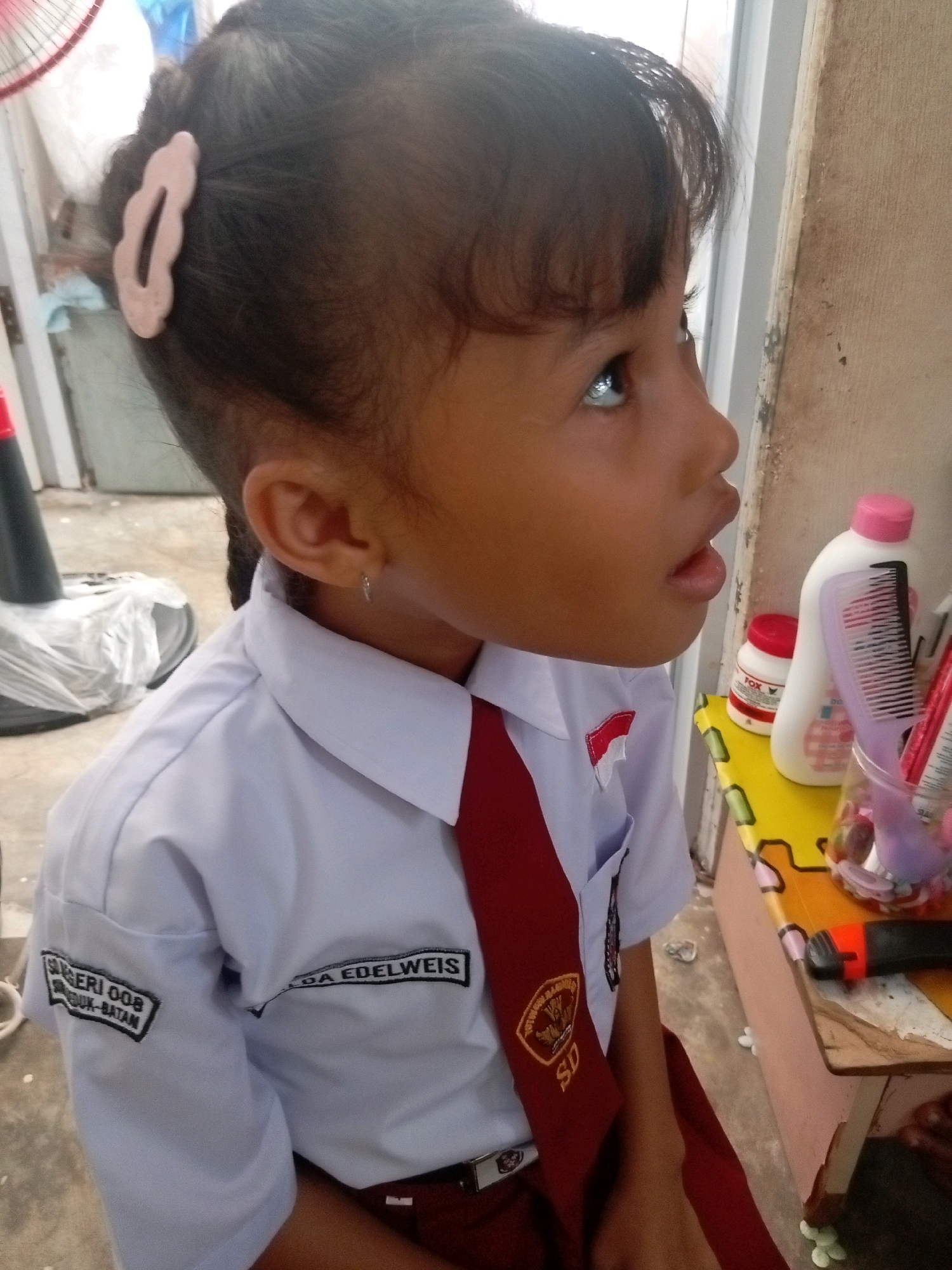 This is a medium shot, eye-level, portrait of a young girl in Indonesia. She is wearing a school uniform with a white collared shirt, a red tie, and a darker red skirt. The shirt has a patch on the left chest with a red and white flag, and another patch on the left sleeve with black text that reads "SUGERI OOS" and below it "DUK-BATAM". The right sleeve has a patch that reads "DA EDELWEIS". She has a pink clip in her hair, which is pulled back into a braid. The girl is looking up and to the right, with her mouth slightly open.
To her right, on a colorful wooden surface, are several objects including a white bottle with a pink lid, a red jar with the word "FOX" visible, a purple comb, and a clear plastic cup filled with small items, possibly candies.
The background appears to be an indoor or semi-outdoor setting with a concrete floor and a light-colored wall. A black traffic cone is visible on the left, and some white plastic sheeting. The lighting suggests it could be daytime, with a slightly overcast or indoor diffused light. The girl's expression is one of curiosity or anticipation.