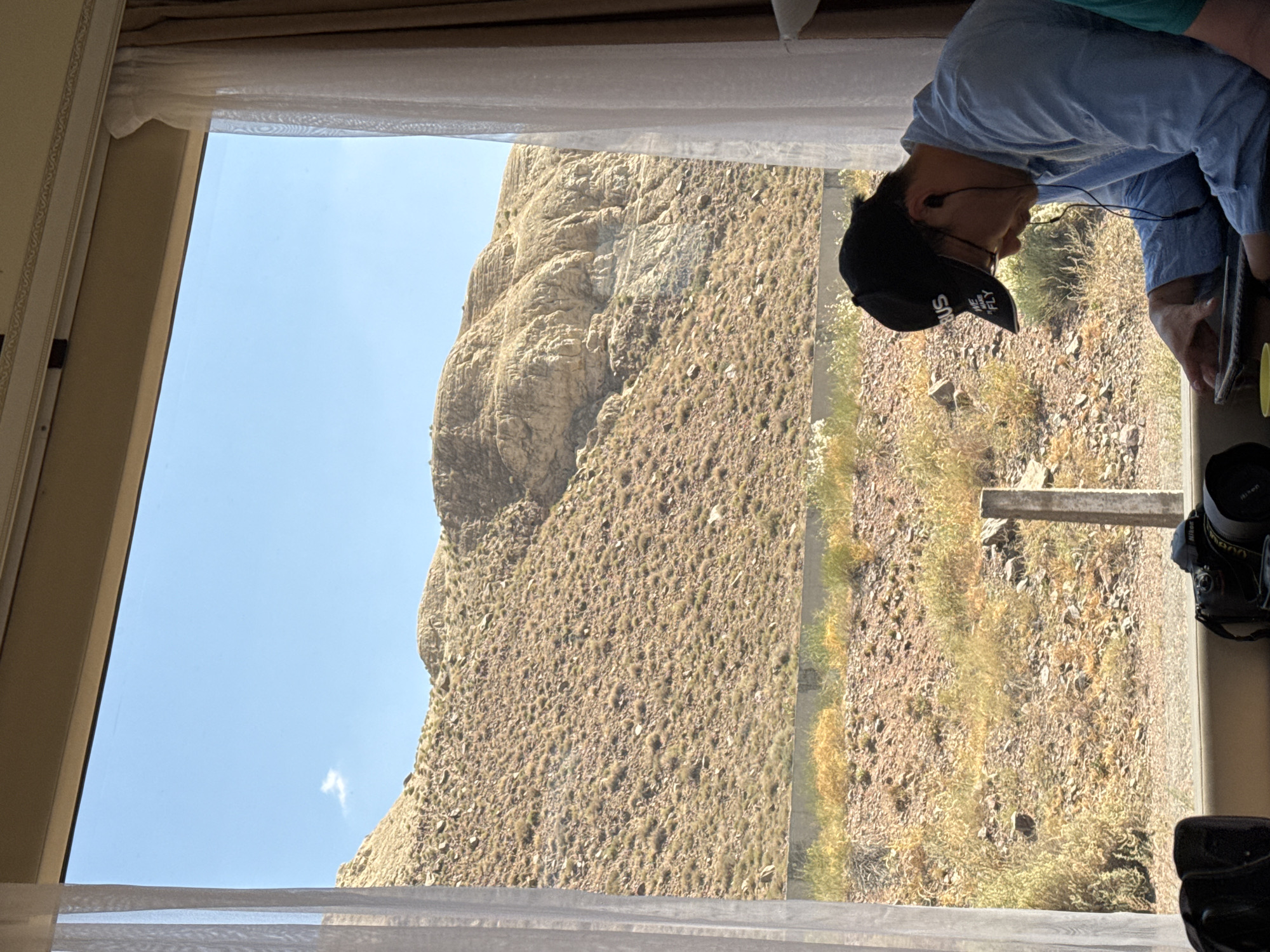 A man wearing a black baseball cap and a light blue collared shirt is looking down at a tablet he is holding in his hands. He has earbuds in his ears. The man is sitting in front of a window with a view of a rocky, arid hillside. Sheer curtains are visible on either side of the window. A camera is on the windowsill in front of him, to his left. To the right of the camera, a yellow cup is visible. The scene appears to be set in a dry, mountainous region.