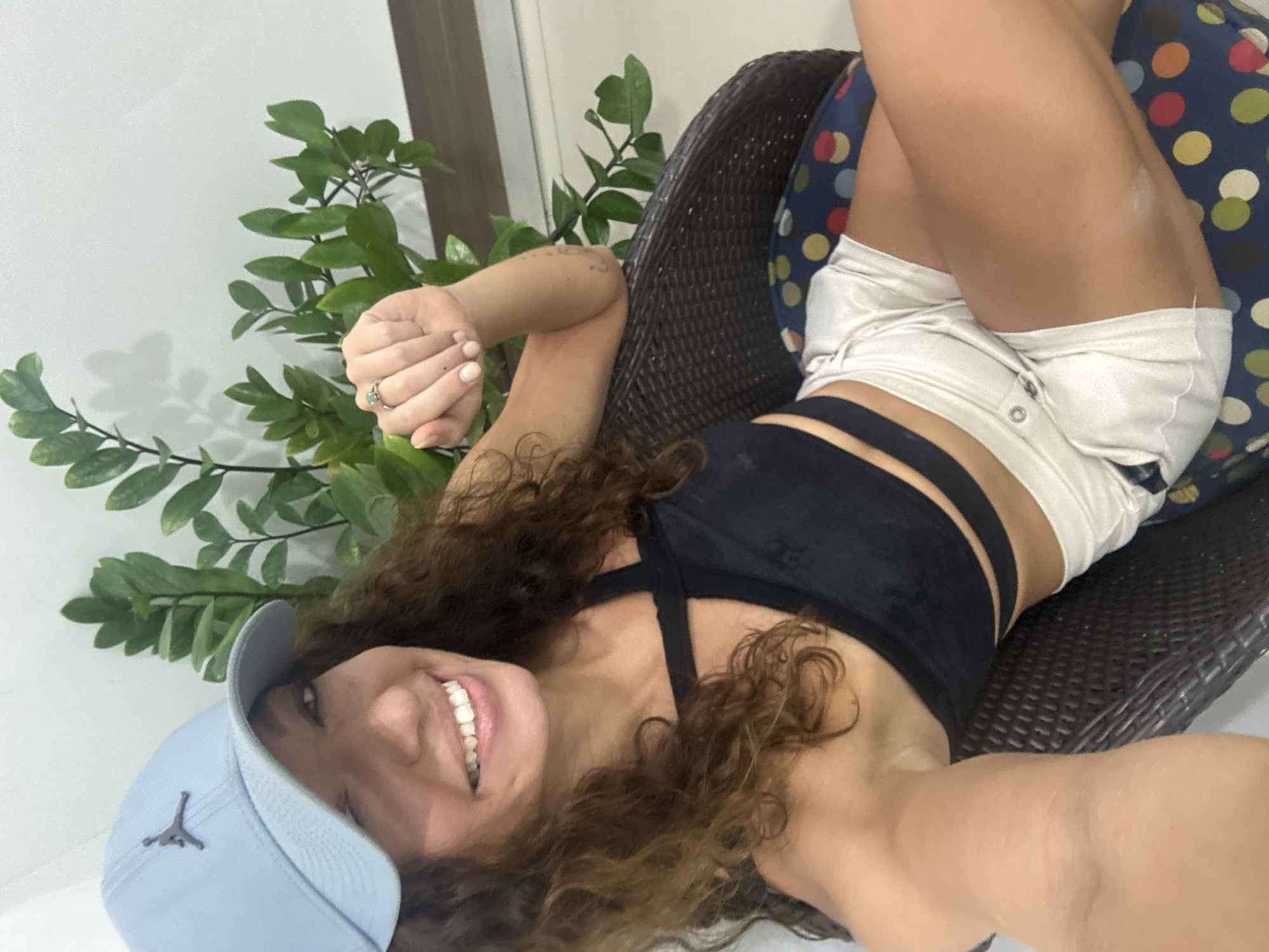 A woman with curly brown hair is smiling at the camera while lying in a dark wicker chair with a polka-dot cushion. She is wearing a light blue baseball cap with a Jordan logo, a black sports bra, and white shorts. To her left is a potted plant with green leaves. The background consists of a white wall and a partially visible doorway. The location is indeterminate from the image.