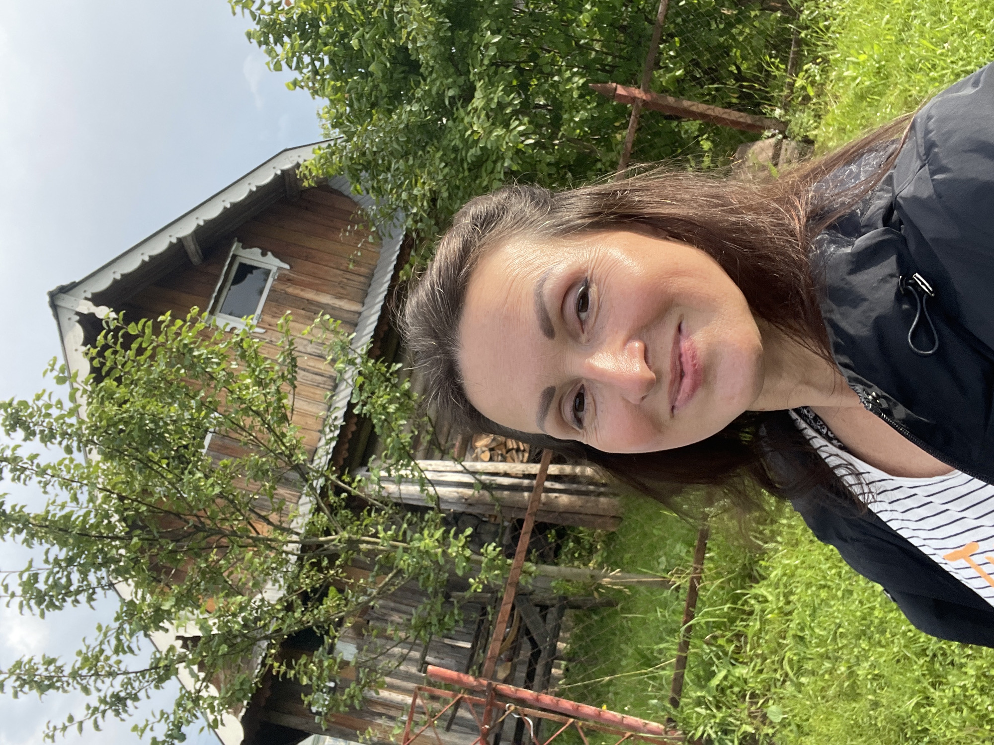 A woman takes a selfie outdoors in a rural setting, with a wooden building and lush greenery in the background. The building appears to be a rustic house or shed, constructed from weathered wooden planks. It has a pitched roof with visible trim detailing and a small window. A tree with dense green foliage stands in front of the building, partially obscuring it. To the left of the building, a red metal fence can be seen. The ground is covered in vibrant green grass, suggesting an outdoor environment. The woman is positioned in the foreground, smiling at the camera. She is wearing a black jacket over a striped top. The sky is partly cloudy. This scene is located in the village of Дресвяново, Russia.