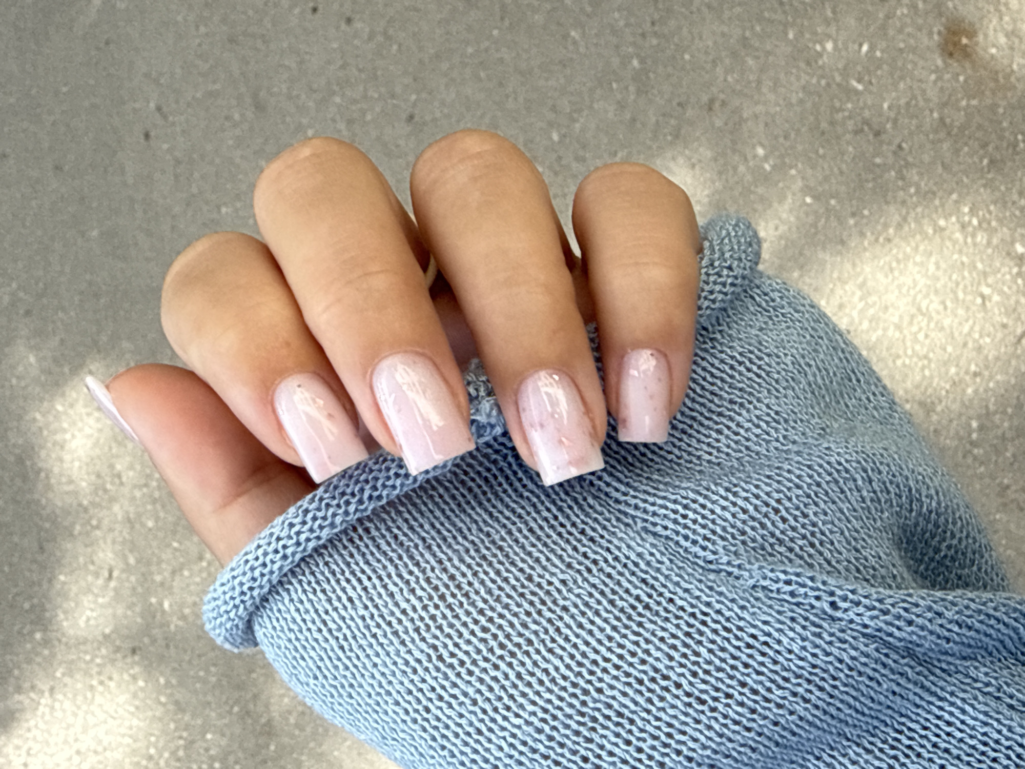 A close-up shot shows a person's hand with light pink, square-shaped nails adorned with subtle glitter. The hand is partially covered by the sleeve of a light blue, loosely woven knit garment. The background consists of a grey, textured surface, possibly concrete, with dappled light and shadows. The image is taken outdoors in an unspecified urban location.