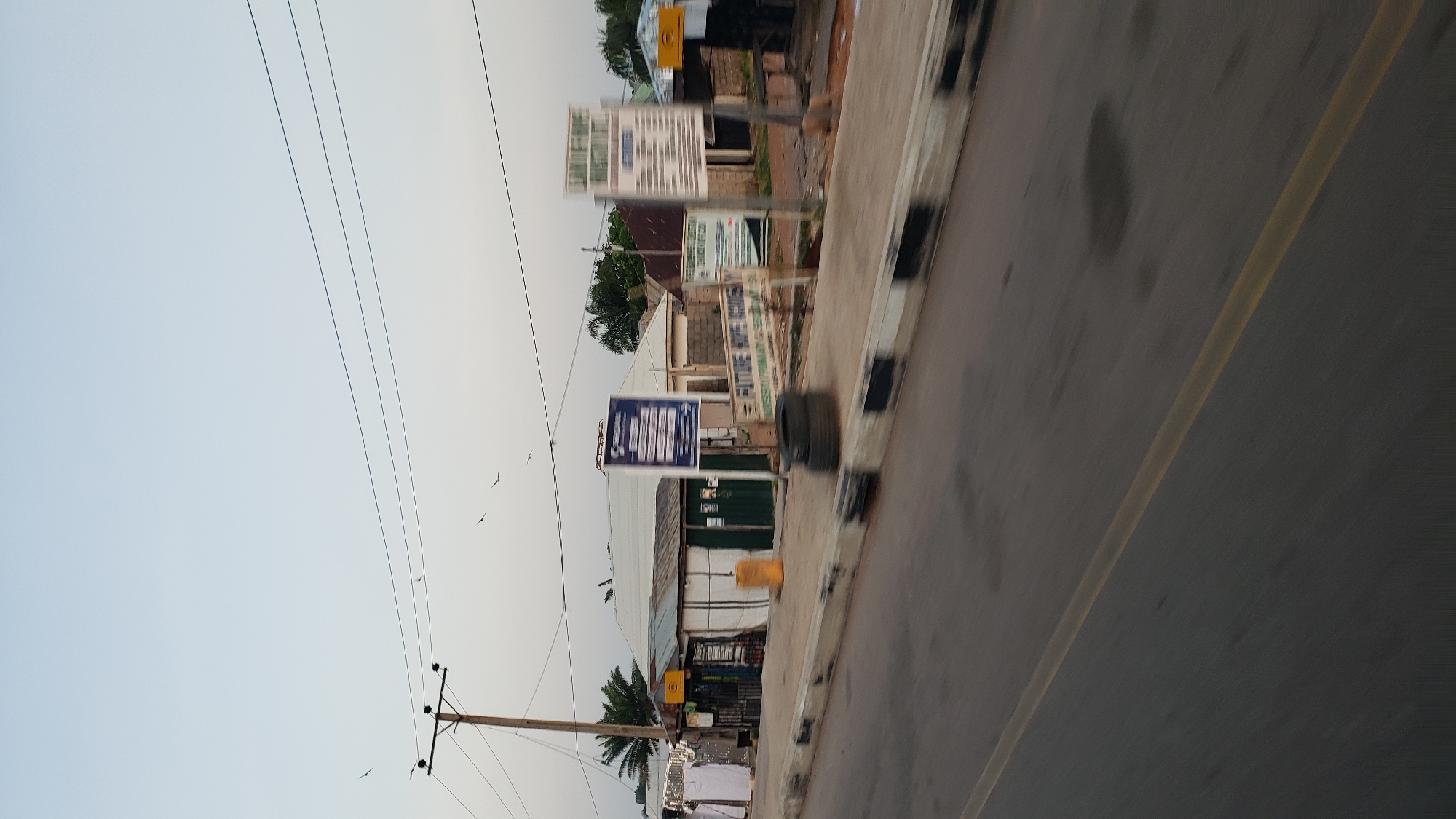 The image captures a street scene in Jalingo, Nigeria, likely during the late afternoon or early evening due to the soft lighting and faint shadows. A paved road with a yellow lane divider occupies the foreground, blurred due to motion. Along the roadside, a concrete barrier with alternating black and white stripes runs parallel to the road. Beyond the barrier is a sidewalk or cleared area where several structures and objects are present.
On the sidewalk, there are a few shops with corrugated metal roofs and open fronts, suggesting they are likely local businesses. One shop has green double doors and is partially covered by a white tarp. Nearby, a yellow cylindrical container is visible. Several white garments are hanging on a line in front of one of the establishments, possibly for sale or drying.
Several signs are placed on poles along the sidewalk. One large blue sign with white text is prominently displayed in front of a shop. Another sign, predominantly white with black text, is further down the sidewalk, and another is partially visible to its right. A smaller sign with "FUTURE HOPE ACADEMY" in large letters is also visible, along with other text below it.
A utility pole with electrical wires stretches across the upper part of the image, with several birds in flight against the pale sky. In the background, more buildings, trees, and palm trees can be seen, indicating a somewhat rural or suburban setting within the city. A stack of tires sits on the sidewalk near the road, and a few other indistinct objects are scattered around. The overall impression is one of a busy, active commercial area, typical of a Nigerian town.