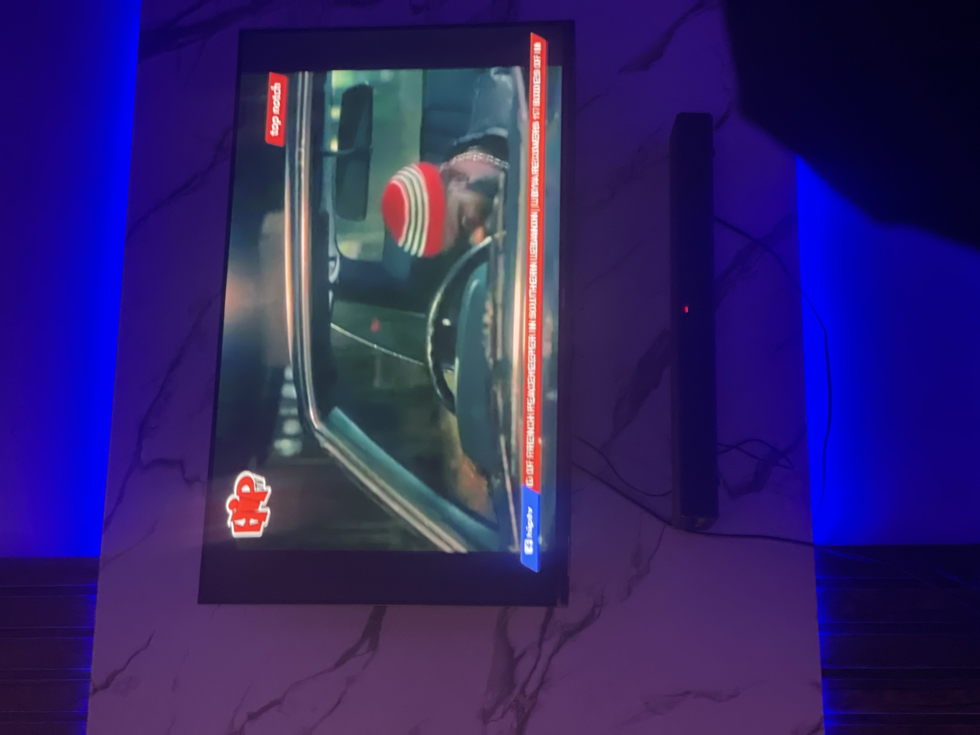 A television screen is displayed on a wall, showing a scene from the Nigerian film "Top Notch," which is currently playing on Iroko TV. The scene appears to feature someone wearing a hat in a car. To the right of the television is a dark, vertical speaker. The room is illuminated with a strong blue light, and the wall behind the screen and speaker has a marbled pattern. The location, Lokoja, Nigeria, cannot be determined from the image.