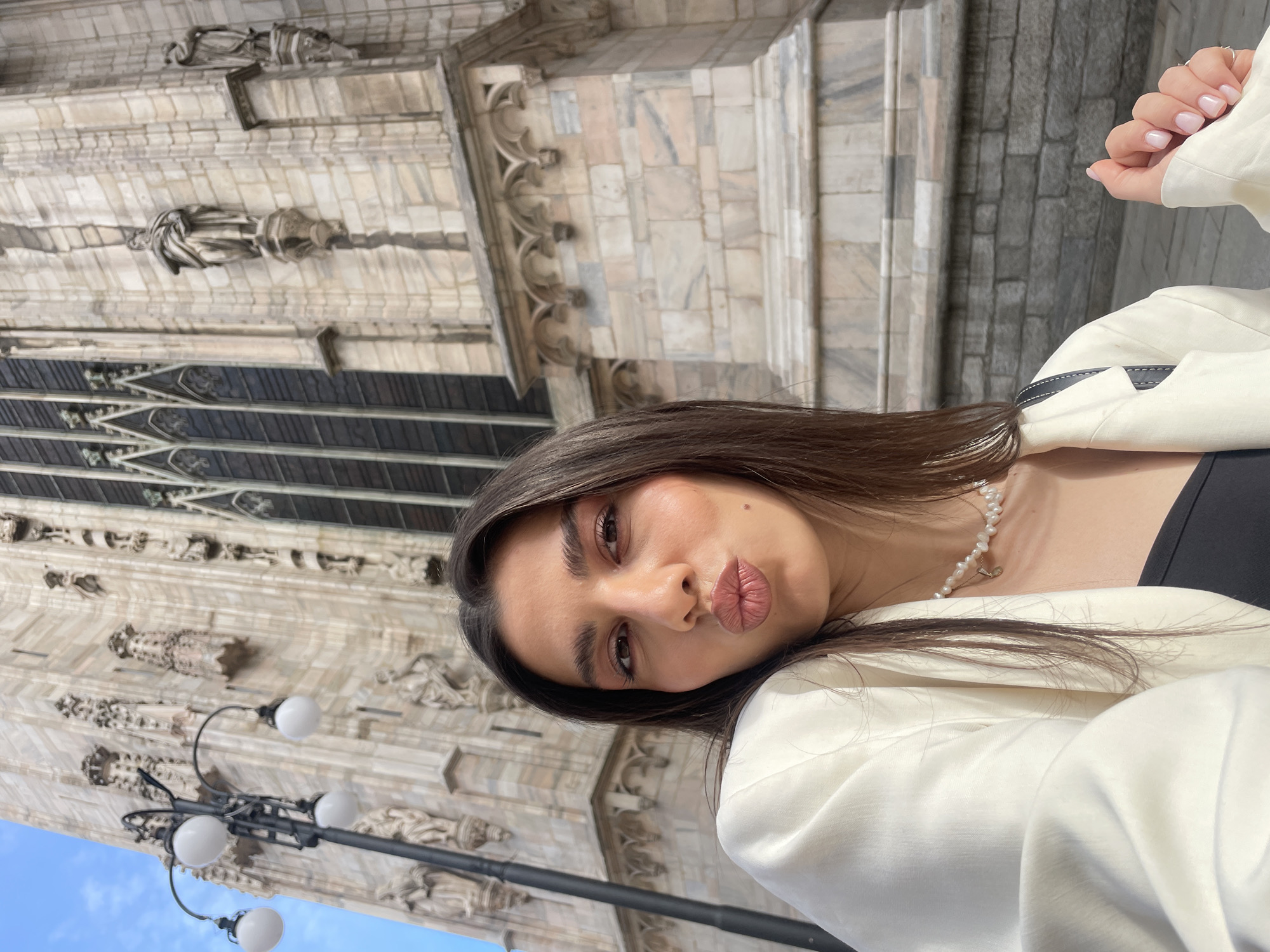 A young woman poses for a selfie in front of the Duomo di Milano in Milan, Italy. She wears a white jacket, a black top, and a pearl necklace. The architecture of the Duomo, including sculptures, windows, and stone construction, is visible in the background, along with a lamppost. The sky is partially visible in the lower left corner.