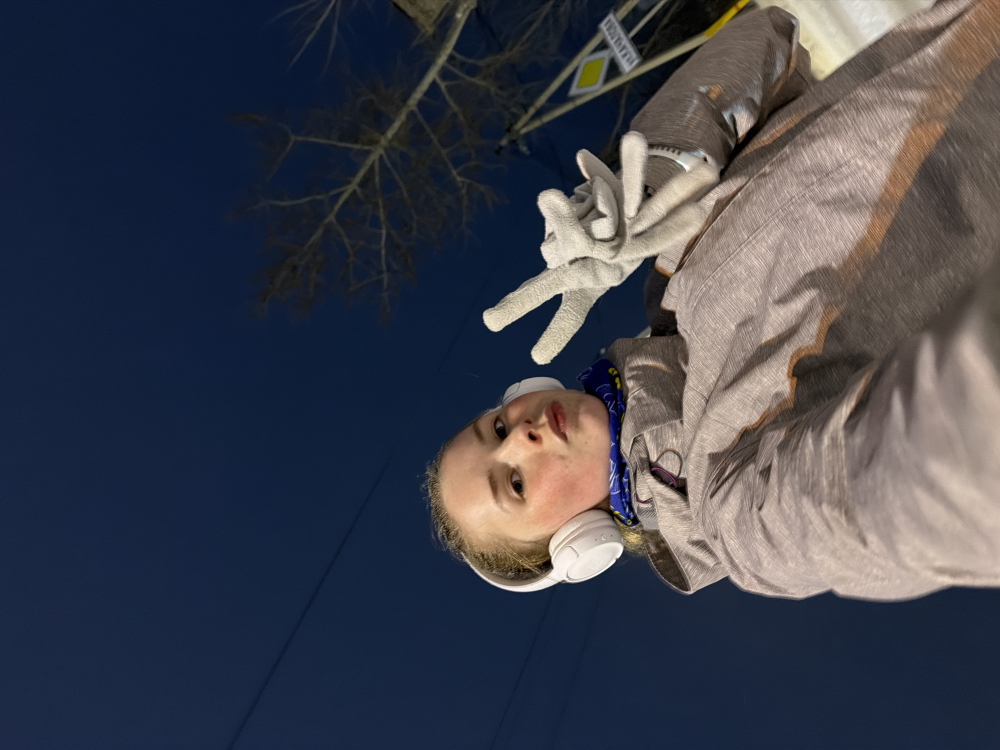 The image captures a selfie of a young person outdoors at night, looking up towards the camera. The individual, appearing to be a young woman with light-colored hair, is positioned in the foreground. She wears large white over-ear headphones, a warm-looking light grey or beige textured winter coat with brown accents, and a blue and white patterned neck gaiter. Her gloved right hand, wearing light grey gloves, is raised to her face, forming a 'peace' sign with her index and middle fingers. Her facial expression is neutral, with slightly parted lips.
The background features a deep, dark blue night sky, indicating evening or night. Silhouetted against the sky on the left are the bare branches of a tree. Towards the right, an urban element is visible: a traffic sign post with two signs attached. The lower sign is a yellow diamond shape with a white border, indicating a 'priority road,' a common sign in Russia. Above it, a white rectangular sign with black text is visible, partially obscured, but the word 'БЕРЕЗНИКИ' (Berezniki) can be discerned, confirming the location as Berezniki, Russia. Faint lines, possibly utility wires or very thin branches, are also visible against the dark sky. The subject's attire suggests cool or cold weather conditions.