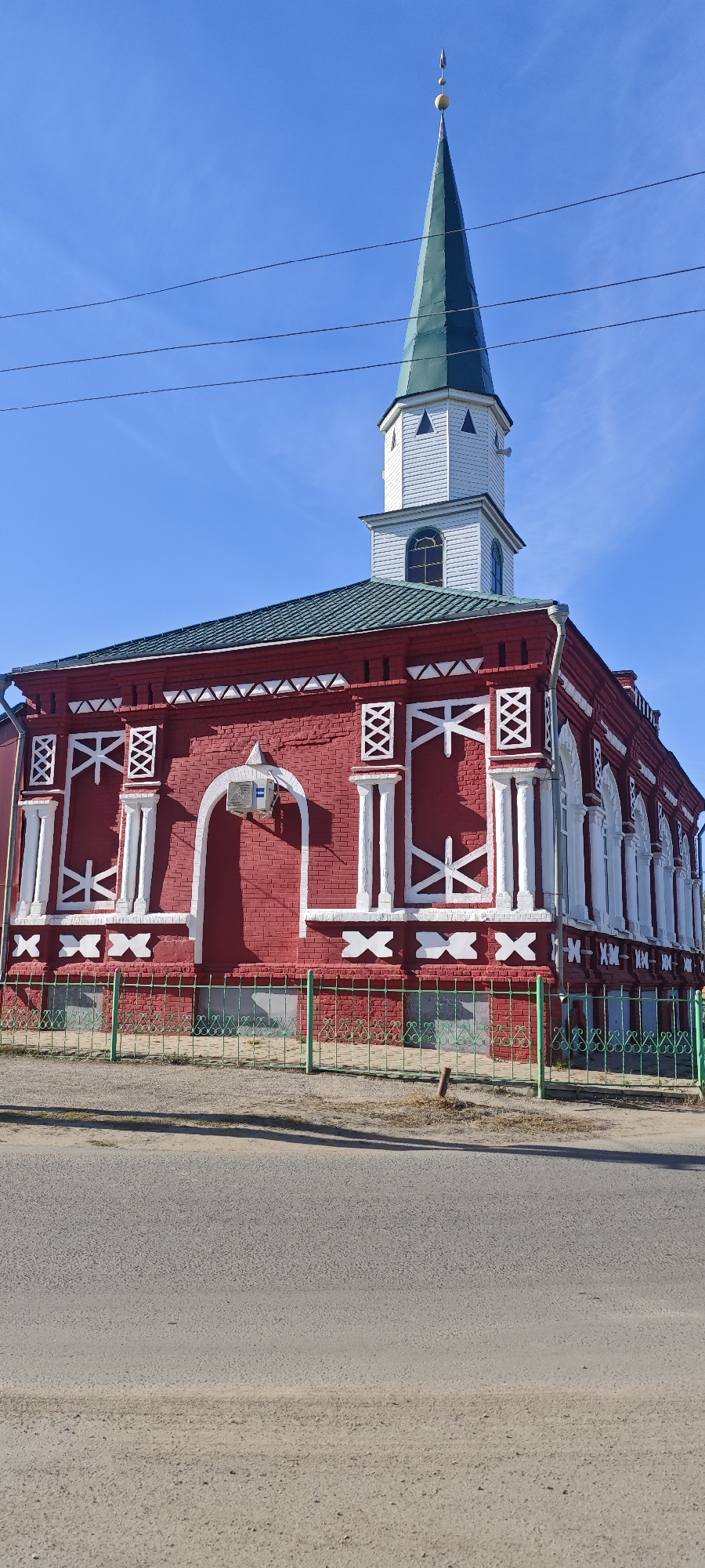 The image depicts the Red Mosque, located in Khvalynsk, Russia, on a clear day with a bright blue sky. The mosque is a two-story red brick building with white decorative elements outlining the corners and windows. It features a green metal roof and a tall, slender minaret with a green spire, mounted on a white base. The mosque is enclosed by a green metal fence. The building faces a paved road. Visible overhead are power lines.