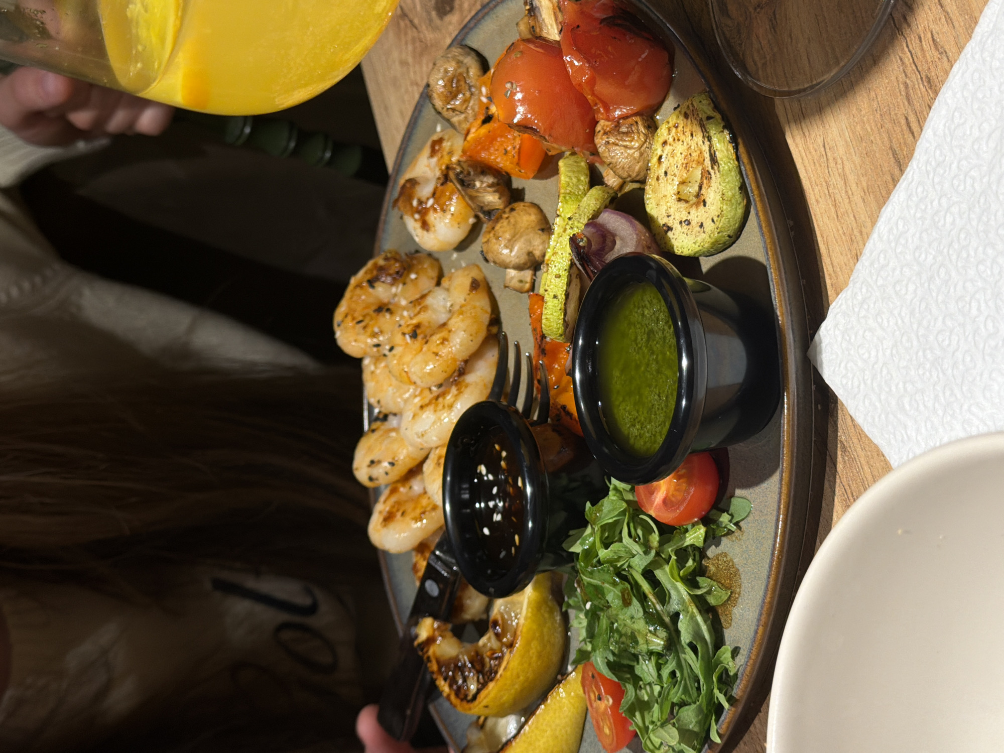 The image shows an oval plate of food, consisting of grilled shrimp sprinkled with sesame seeds, grilled vegetables (tomatoes, mushrooms, zucchini, red onion, and carrots), two small black bowls, green leafy vegetables (arugula), cherry tomatoes, and grilled lemon wedges. One bowl contains a green sauce, and the other contains a dark, possibly soy-based, sauce also sprinkled with sesame seeds. A person's hand is visible holding a knife and fork that are on the plate. A hand is also visible pouring a yellow liquid, possibly juice, from a clear glass. The plate sits on a wooden table next to a white napkin and a white bowl. The background suggests a dimly lit restaurant or dining area. There is no verifiable information to confirm that the image was taken in Chaikovsky, Russia.