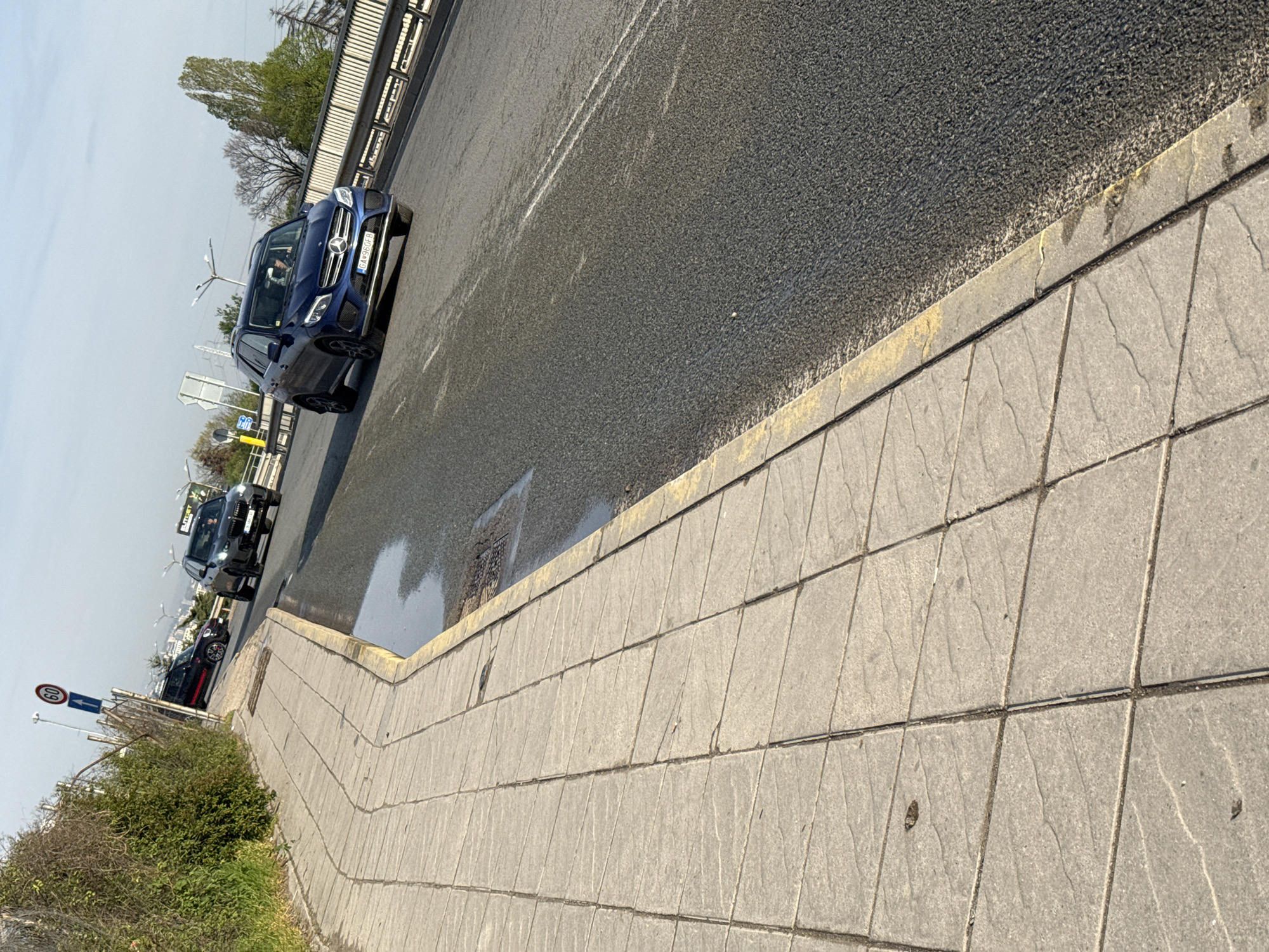 The image captures a tilted, bright daytime scene of a road and adjacent sidewalk, likely in an urban or suburban setting, consistent with Sofia, Bulgaria. The perspective is from the sidewalk looking across the road, with the horizon tilted clockwise.
In the foreground, a gray paved sidewalk with large rectangular stones runs along the right edge, meeting a yellow-painted curb that borders the asphalt road. The road surface appears dark and speckled, with some visible wet patches.
Several cars are on the road. Most prominently, a dark blue Mercedes-Benz SUV (possibly a GLE model) is visible in the mid-ground, heading away from the viewer. Its front license plate, "GA R860FB", is clearly legible, indicating a Bulgarian registration. A person is faintly visible in the driver's seat. Further down the road, another dark SUV (possibly a BMW X-series) is visible, followed by a smaller dark car, possibly a MINI Cooper, with red accents.
To the left of the road, a metal barrier or fence runs along the roadside, and behind it, green foliage from bushes and trees can be seen. Some bare trees suggest it might be early spring. Utility poles or streetlights are visible in the background, and distant buildings hint at a city. A circular speed limit sign displaying "60" (km/h) is present on the left side of the road. A partially visible billboard with the text "BETSET" can also be seen in the distance. The sky above is light blue with some haze. The bright sunlight suggests the image was taken during clear weather conditions.