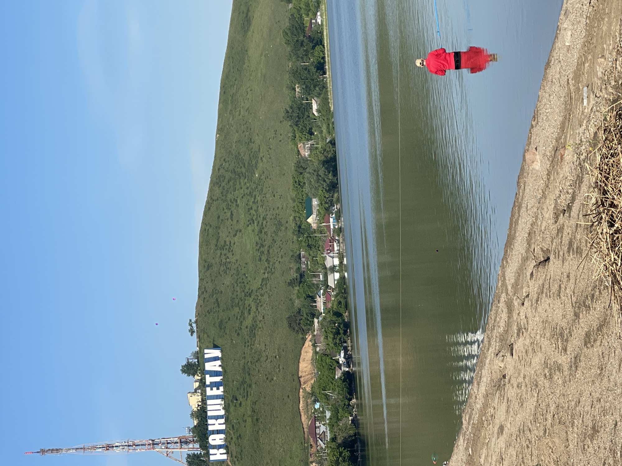 The image shows a scene in Kokshetau, Kazakhstan. A body (possibly a doll) dressed in red is floating in the water. The water reflects the light. A concrete structure is visible on the right side of the image. In the background, there is a hill covered in greenery, with buildings and houses scattered along its base. The "KOKSHETAU" sign is visible on the hillside. There is a communications tower in the background. The sky is blue.