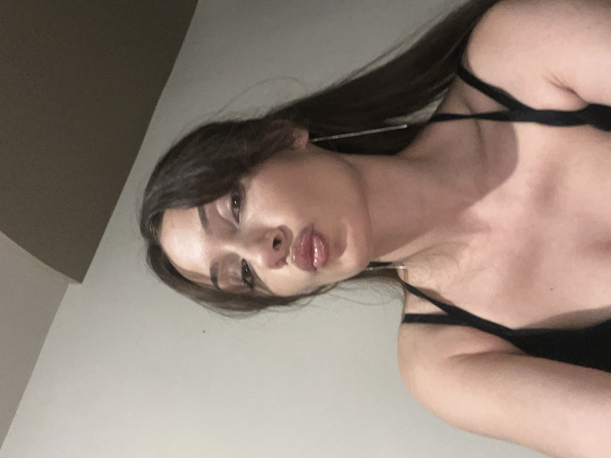 A medium-shot of a woman is shown at a slight angle. The woman has long, dark brown hair that cascades over her left shoulder. She is wearing a black, thin-strapped top. Her face is angled towards the camera, and she is puckering her lips. She is wearing long, silver earrings. The background is a plain, light gray wall with a corner of a darker gray wall visible in the upper left. There is no indication of the location being in Burgas, Bulgaria.