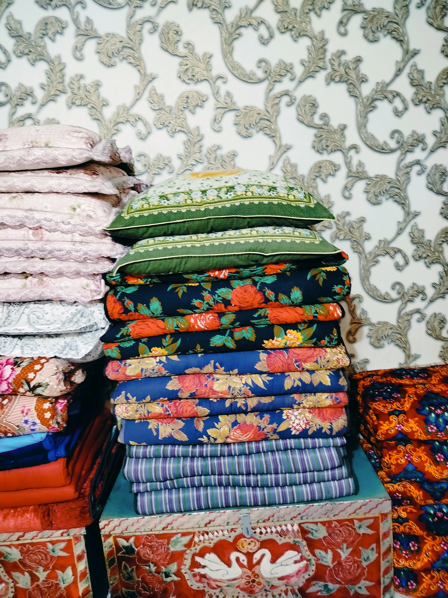 The image displays a stack of colorful folded textiles and pillows against a patterned wallpaper. The stack on the left consists of pink and white pillows with delicate lace trim, followed by white and grey patterned pillows. To the right, a stack of pillows with green floral patterns tops a larger pile of folded fabric. This lower stack features fabric with bold floral designs in red, orange, and yellow on dark blue backgrounds, transitioning to a blue fabric with pink and yellow floral patterns, and finally a stack of striped fabric in shades of purple and green.
In the foreground, to the left, are stacks of folded red and orange fabric, some with floral patterns. To the right, a colorful, patterned textile with orange, blue, and black elements is visible. The bottom of the image shows ornate decorative boxes, one with a red floral pattern and two white swans, and another with red floral designs. The wallpaper behind the textiles features a repeating ornate, scrolling pattern in light grey on a white background. The overall scene suggests an indoor setting, possibly a storage area or display of textiles. The specific location in the city of Көлкент, Kazakhstan is not determinable from the visual information provided.