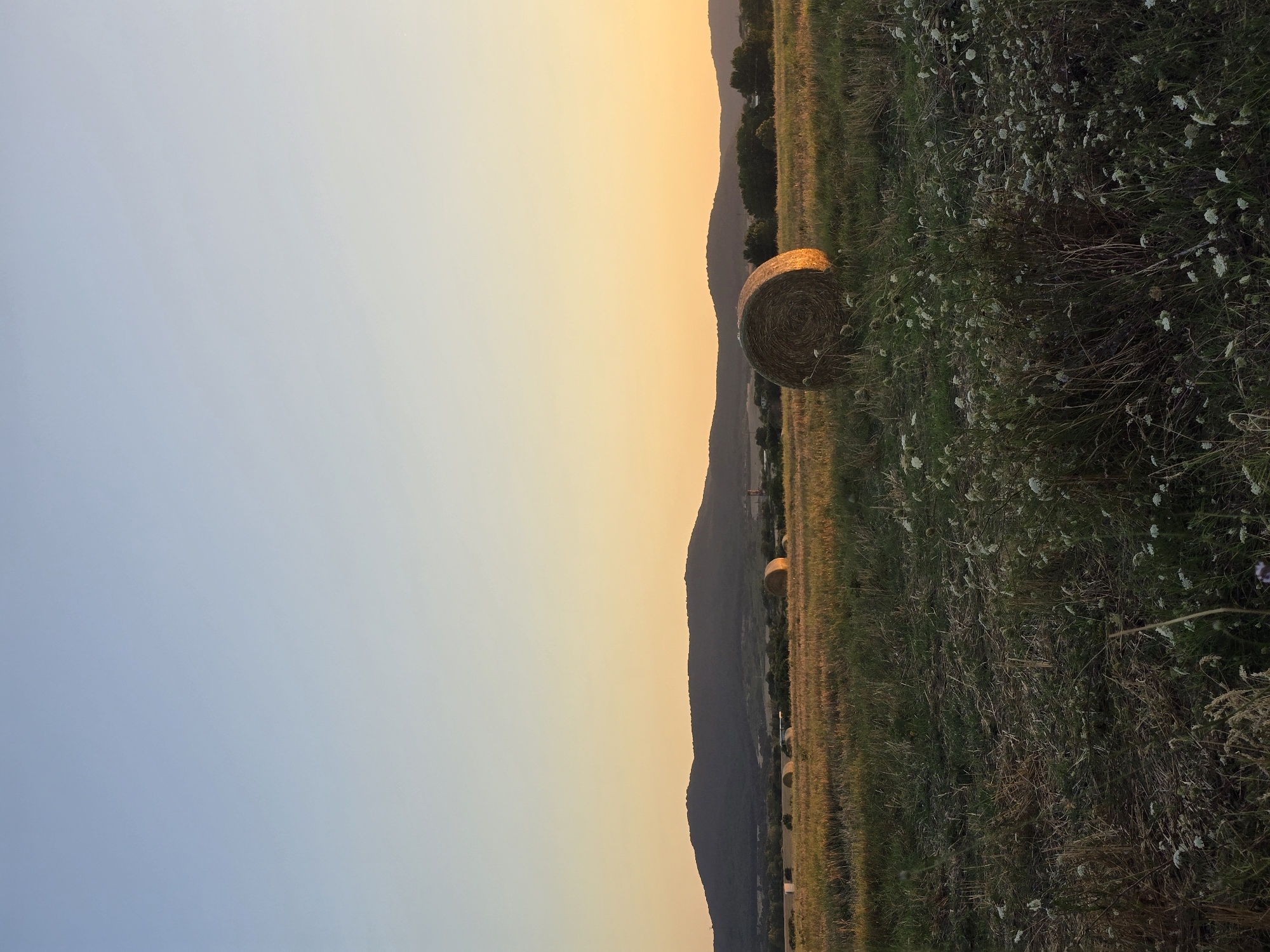 The image depicts a rural landscape at sunset near Chernomorets, Bulgaria. In the foreground, a field of dry grass and wildflowers is visible, with a large, round hay bale prominently positioned in the middle ground. The hay bale is illuminated by the warm glow of the setting sun, casting a golden light on its surface. In the background, a range of hills forms a dark silhouette against the vibrant orange and yellow hues of the sunset sky. Several other hay bales and scattered trees are also visible in the distance, contributing to the pastoral scene. The overall impression is one of tranquility and the beauty of nature at dusk.