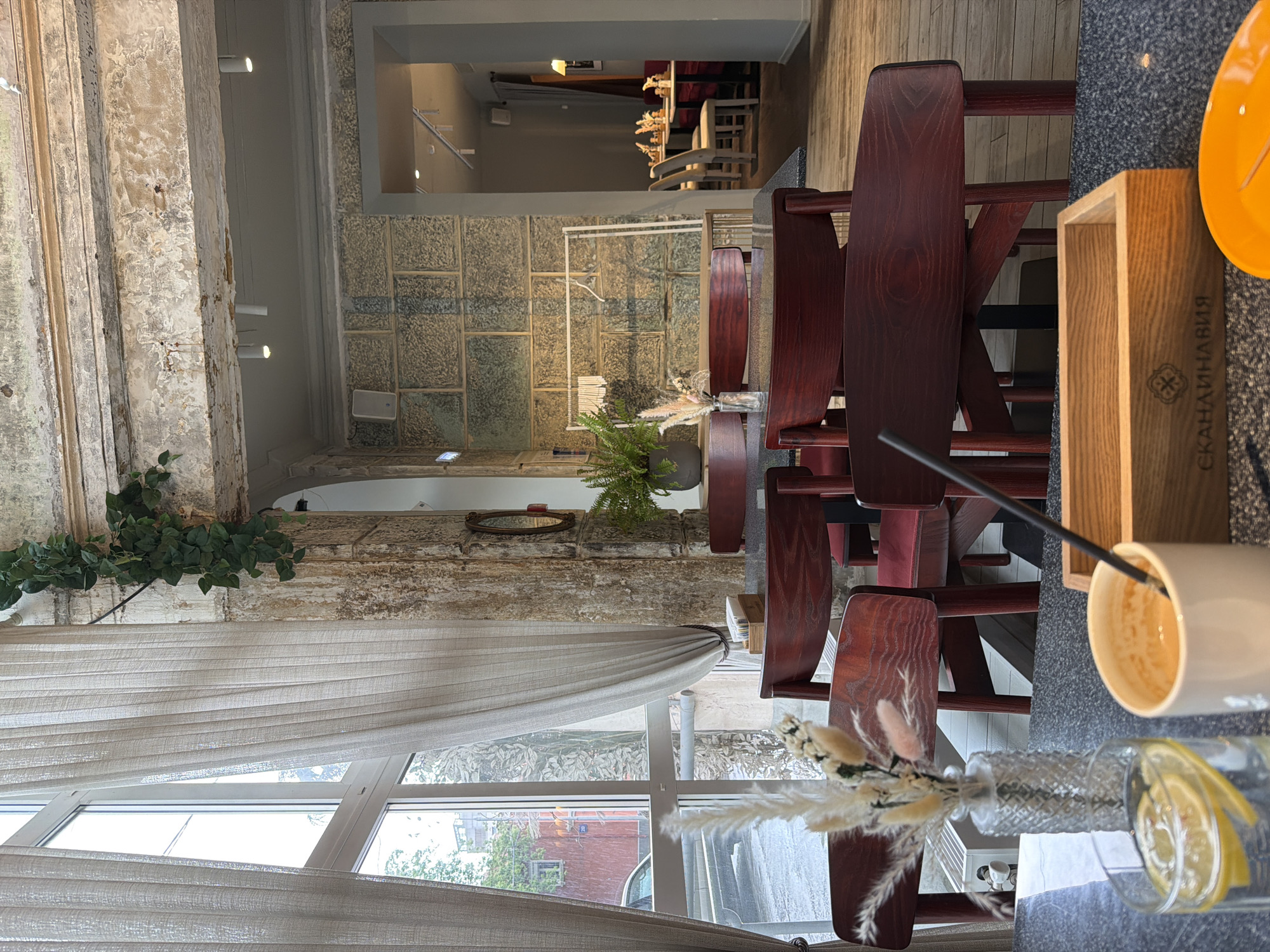 The image shows the interior of a café in Перовское сельское поселение, Ukraine. The café features rustic, textured walls and a large window with a sheer curtain. In the foreground, there's a table set with a glass of water with a lemon slice, a coffee cup, and a wooden container. The table is surrounded by dark red wooden chairs. There is a vase with dried flowers on the table. A plant in a pot sits further back on the table. Through a rectangular opening, another area of the café can be seen with additional tables and chairs.
